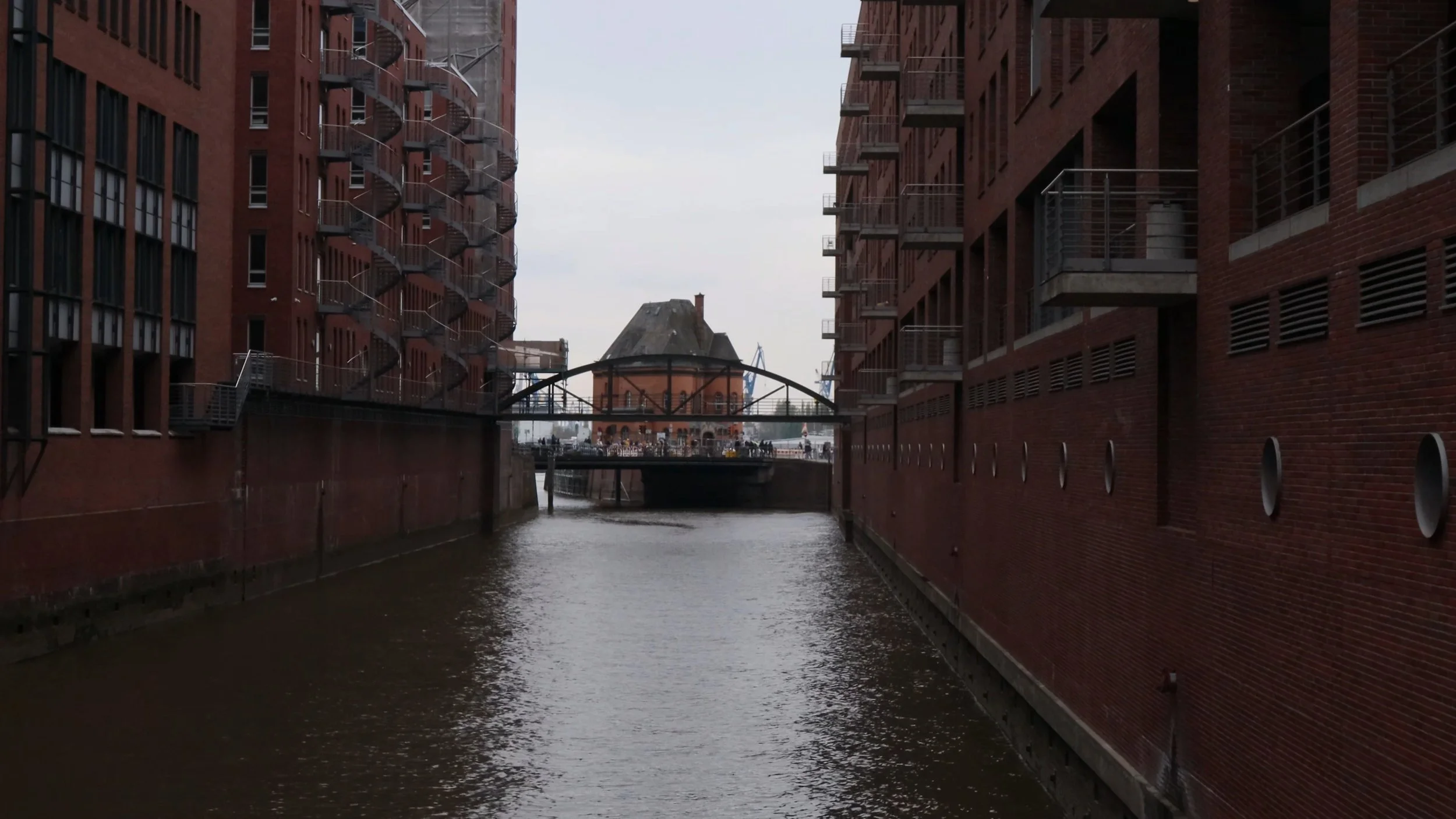 View of a narrow canal flanked by red brick apartment buildings with fire escape stairs, leading to a bridge and historic brick building in the distance.
