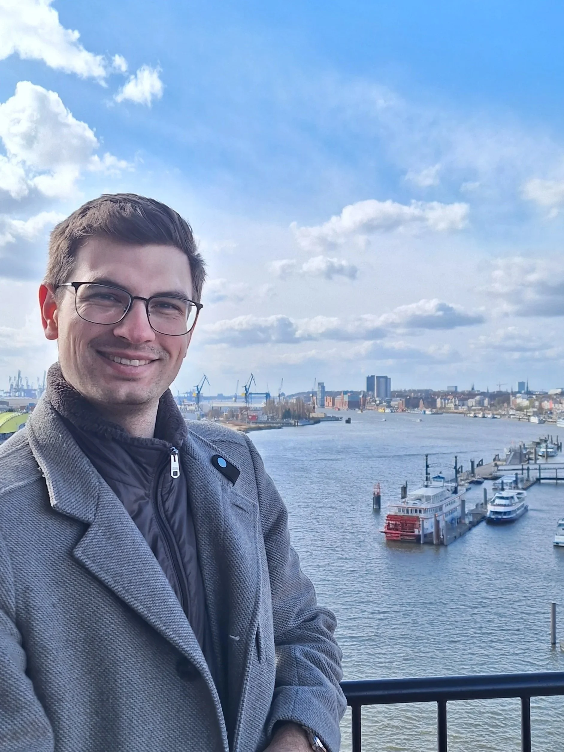 A man wearing glasses and a gray coat smiling at the camera, with a river, boats, cranes, and city buildings in the background under a partly cloudy sky.