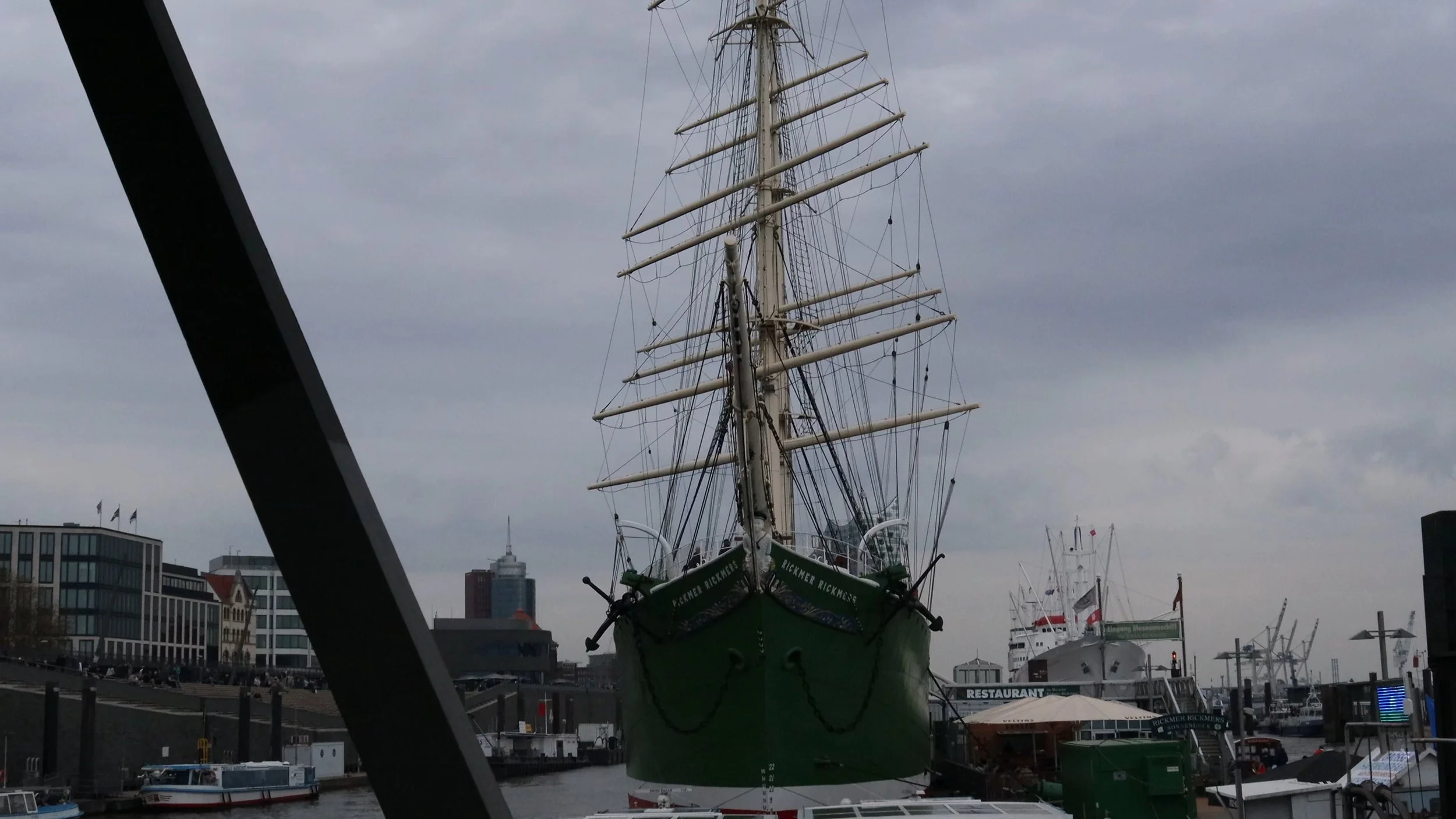 A large sailing ship docked in a harbor with modern buildings in the background. The ship has multiple masts and rigging.