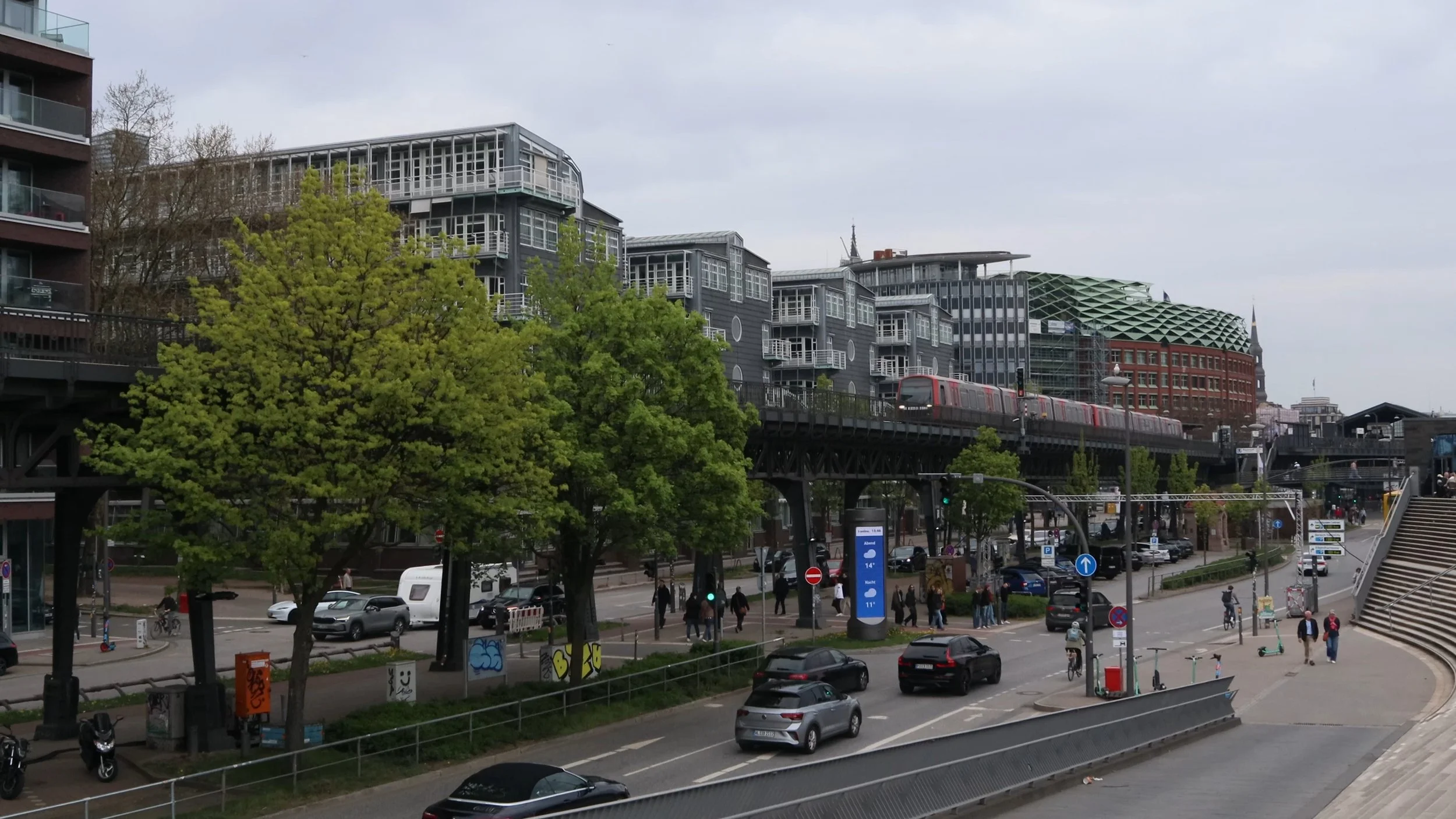 Urban city scene with modern buildings, a train on elevated tracks, cars on the street, pedestrians, trees, and stairs, overcast sky.
