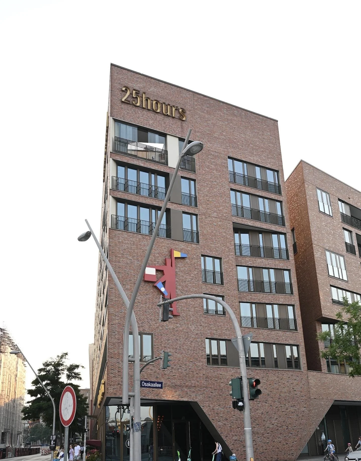 A tall brick building with multiple floors, large windows, and balconies. A street sign reads 'Osakallelée.' There are streetlights, traffic lights, and pedestrians, including cyclists, at the street level.