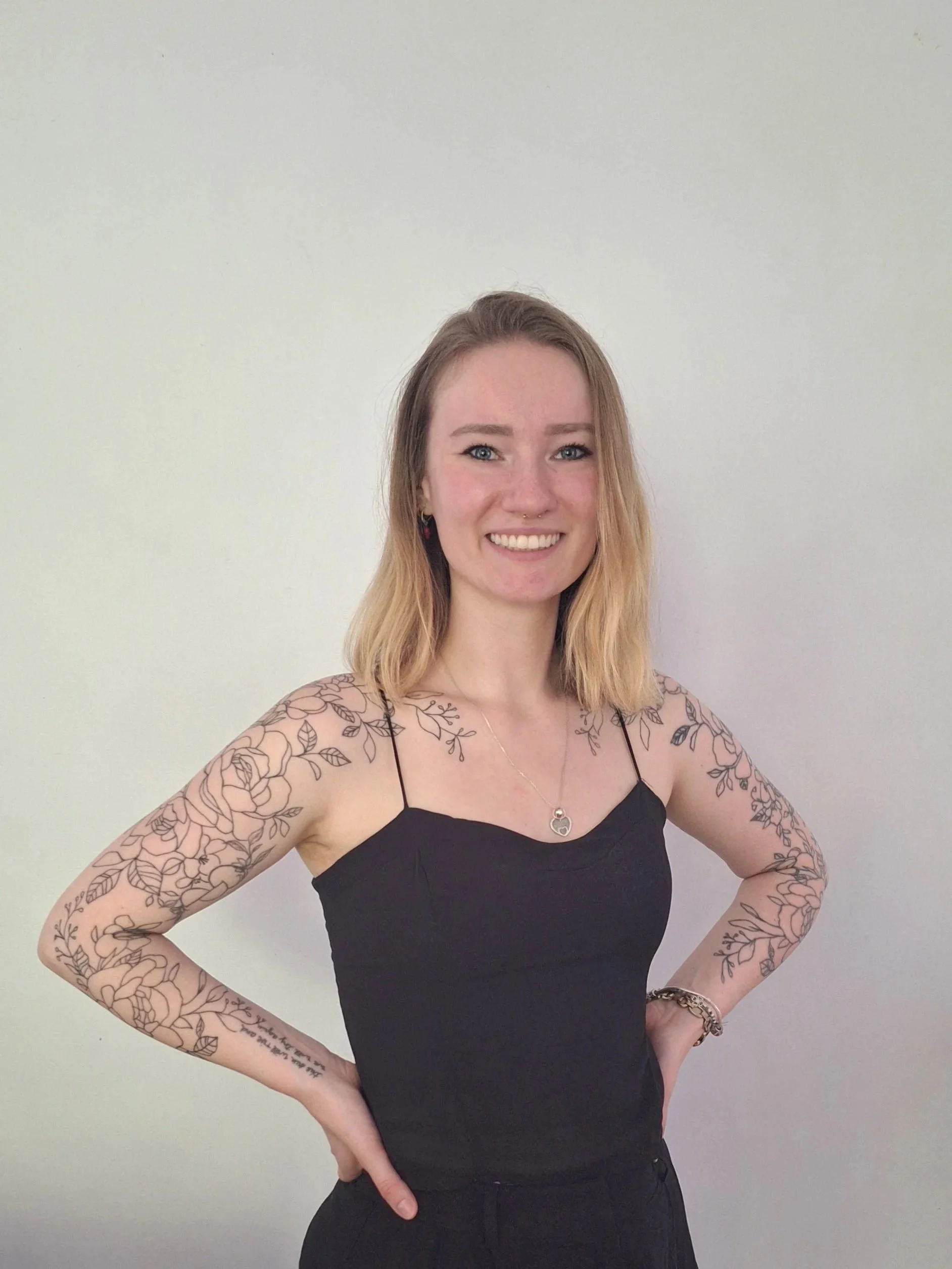 A woman with shoulder-length blonde hair, blue eyes, and tattoos on her arms standing against a plain white wall. She is smiling, wearing a black sleeveless top and jewelry, with her hands on her hips.
