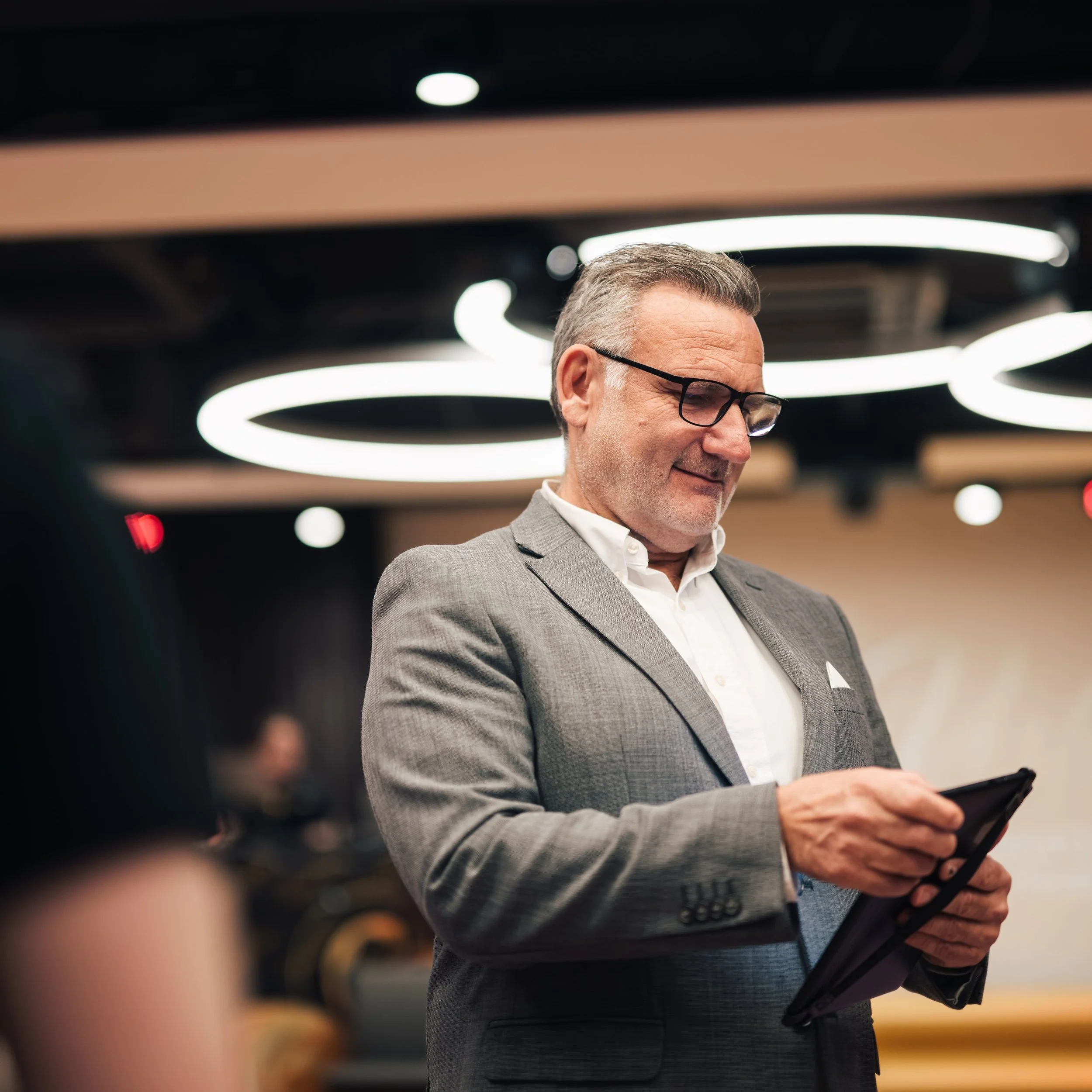 A middle-aged man with gray hair, wearing glasses and a gray suit, is looking at a smartphone. The background features a modern ceiling with circular light fixtures.