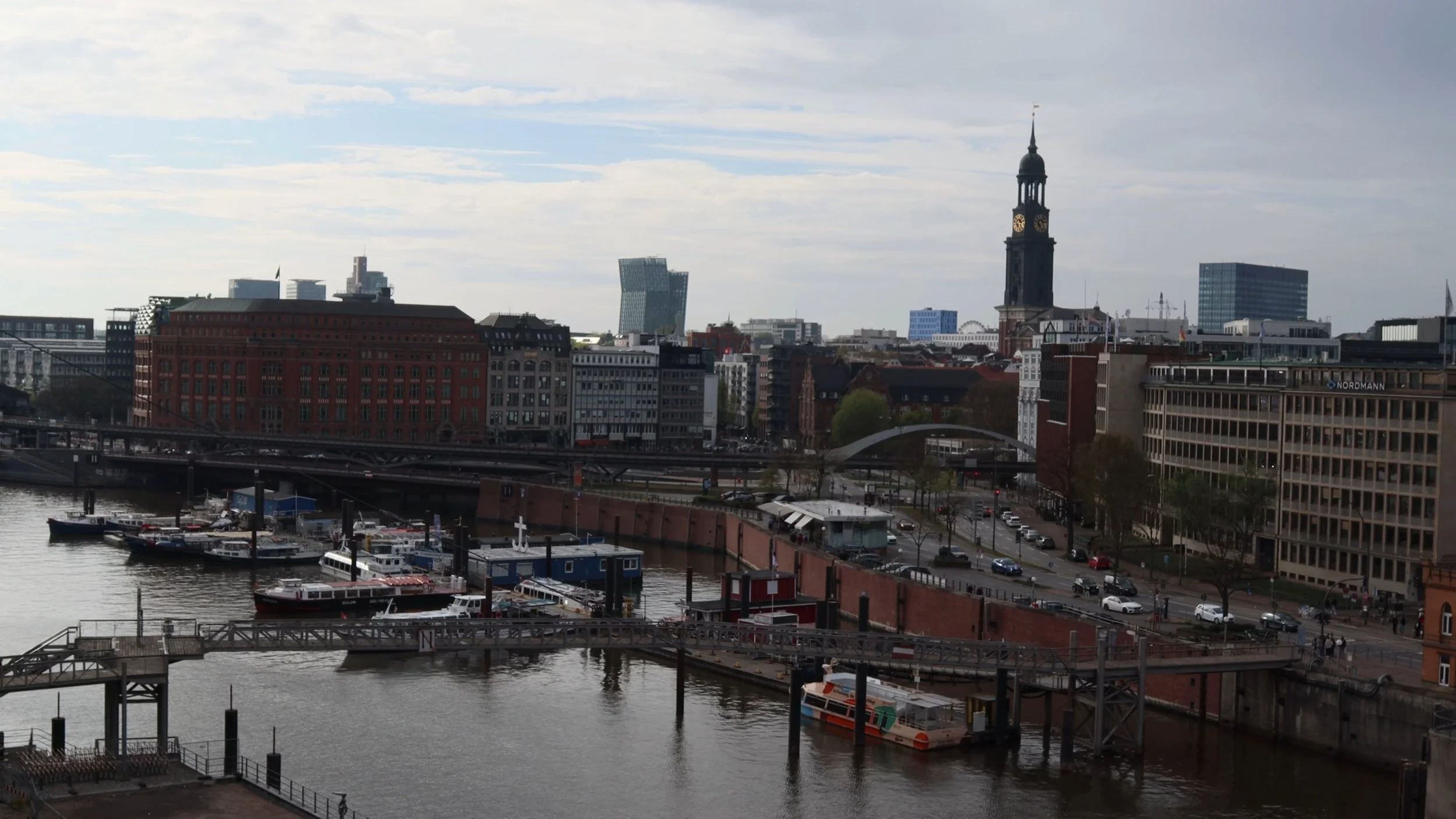 View of a city skyline including historic and modern buildings, a church tower, a bridge over a river, and boats docked at a harbor on a cloudy day.