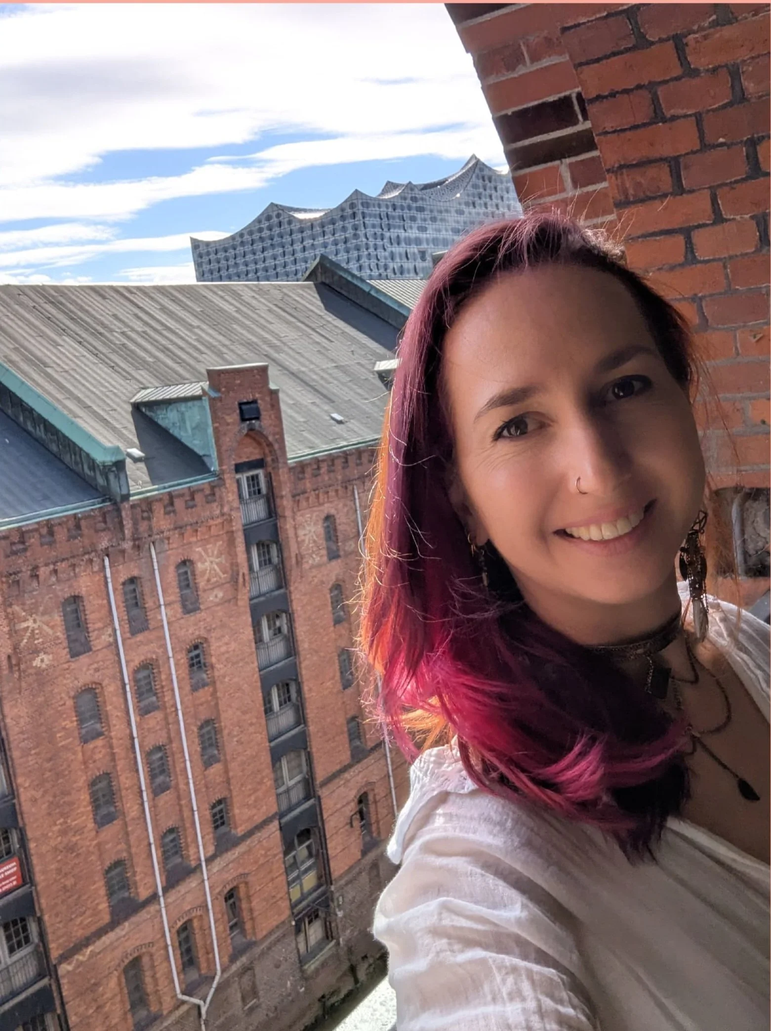 A woman with pink hair smiling on a balcony with a brick wall. In the background, there are old brick buildings and a modern building with a wavy glass roof.