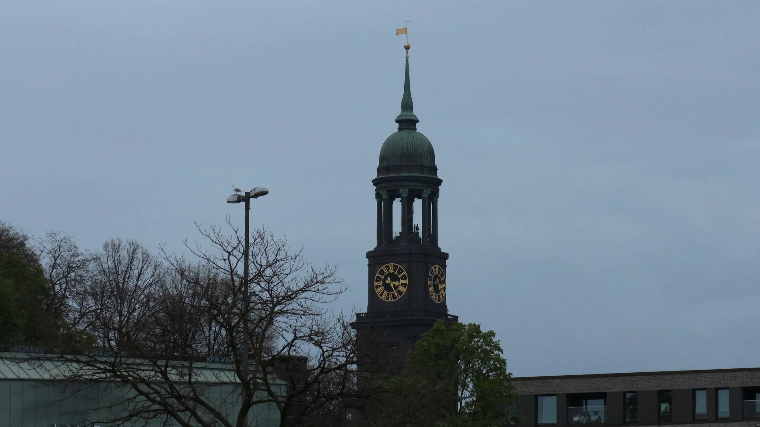 Clock tower with a green dome and golden weather vane, trees, and a building in the foreground during daytime.