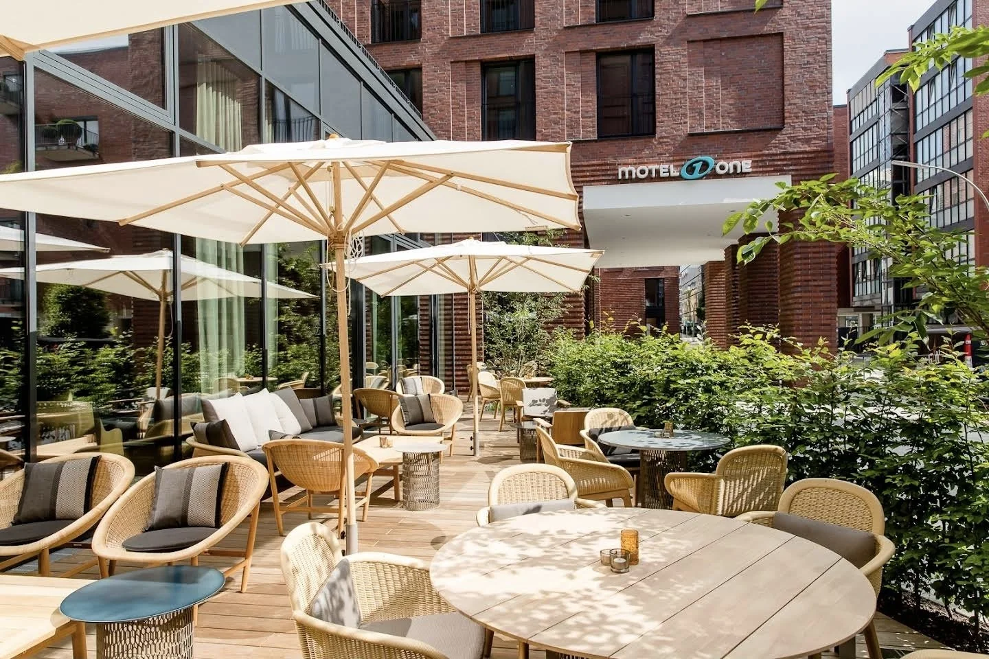 Outdoor patio with wicker chairs, tables, and large umbrellas outside a hotel or restaurant, with bushes and a brick building in the background.