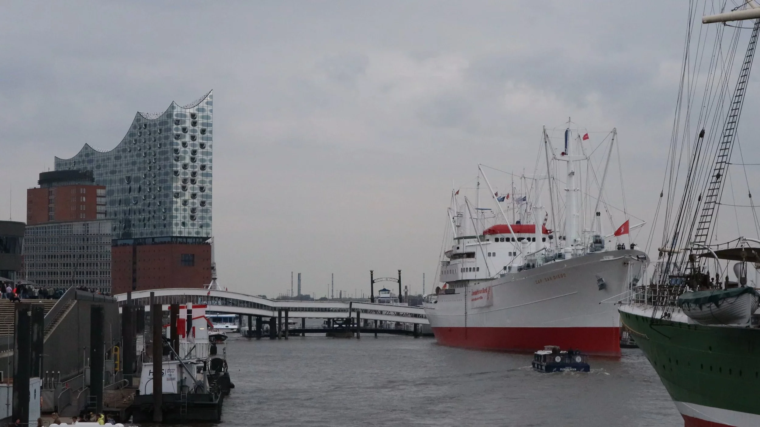 A harbor scene with boats docked at a marina, including a large white yacht with a red stripe, and a modern glass building with a wavy design in the background under a cloudy sky.
