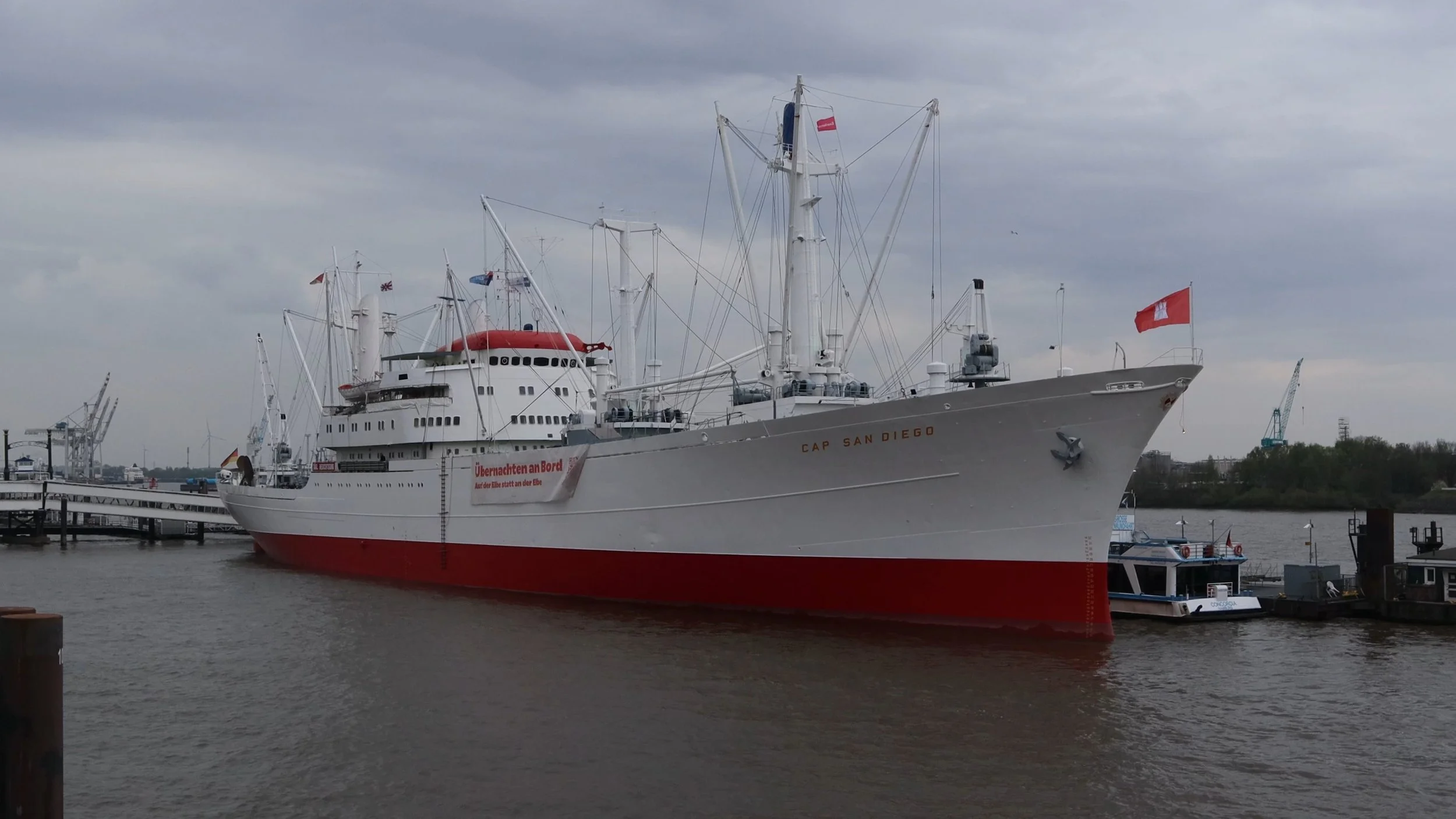 Large white and red cruise ship named 'Cap San Diego' docked at a harbor with cranes in the background and a cloudy sky overhead.