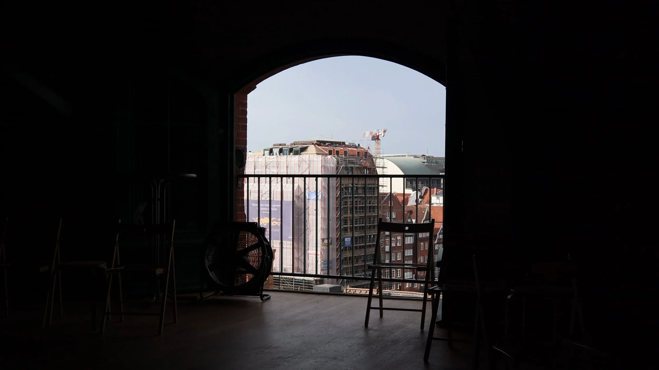 Cityscape view through a large arched window from inside a dark room, with chairs and a fan in the foreground.