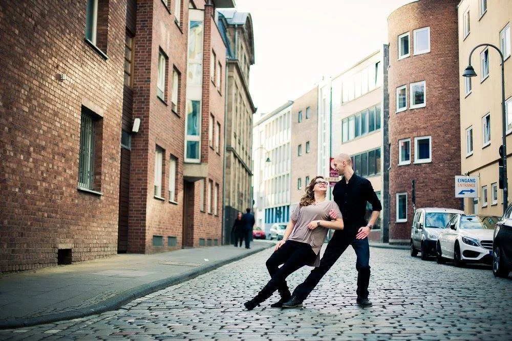 A man and woman dancing on a cobblestone street in an urban setting, surrounded by modern buildings and parked cars, with a sign indicating the entrance in German.