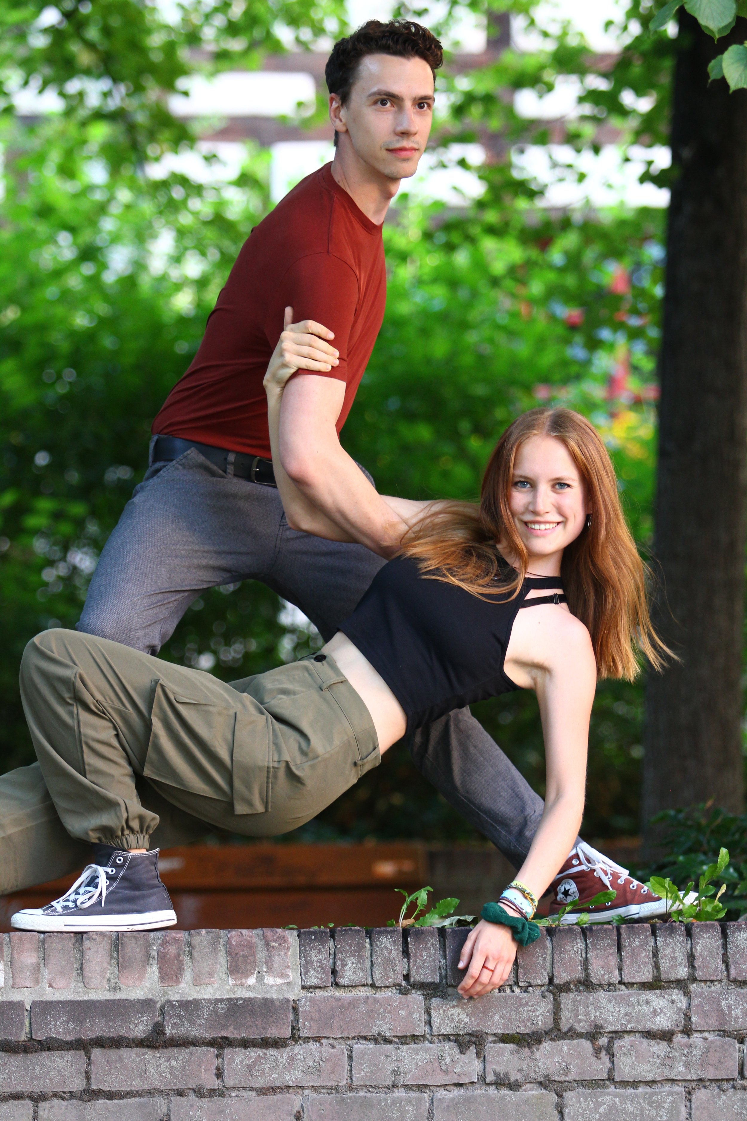 Two young adults, a man and a woman, are posing outdoors on a brick ledge with trees and greenery in the background. The man is standing behind the woman, who is crouching on the ledge, and they are holding hands in a supportive pose.