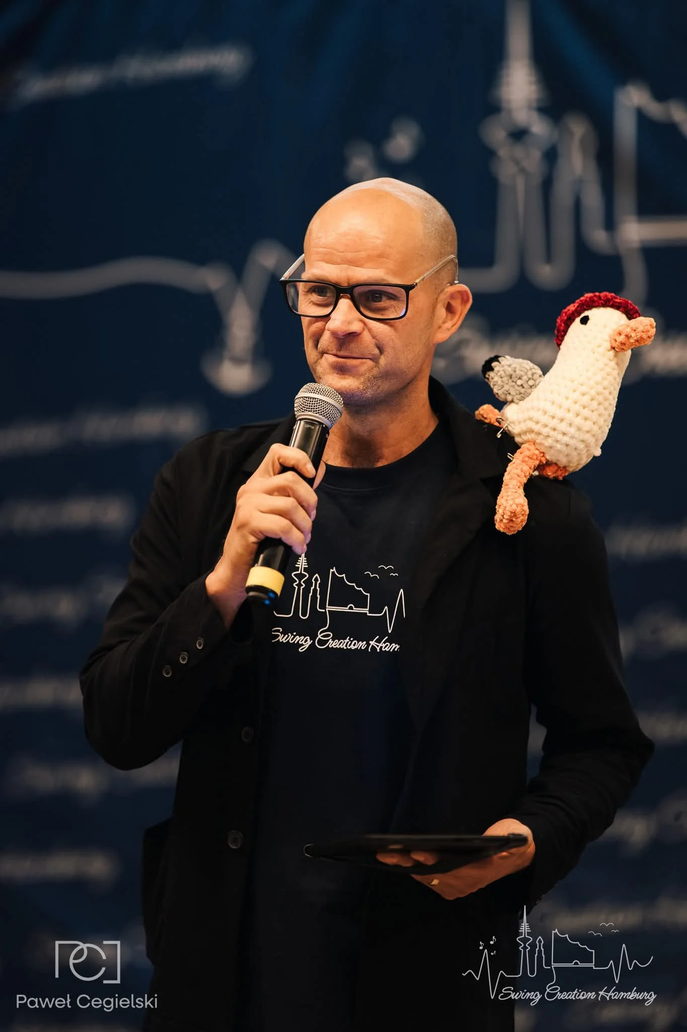 A man wearing glasses and a black cap holds a smartphone with a patterned strap close to his ear, smiling slightly. There is a woman in the background and a microphone in front of him, indicating a public or media event.