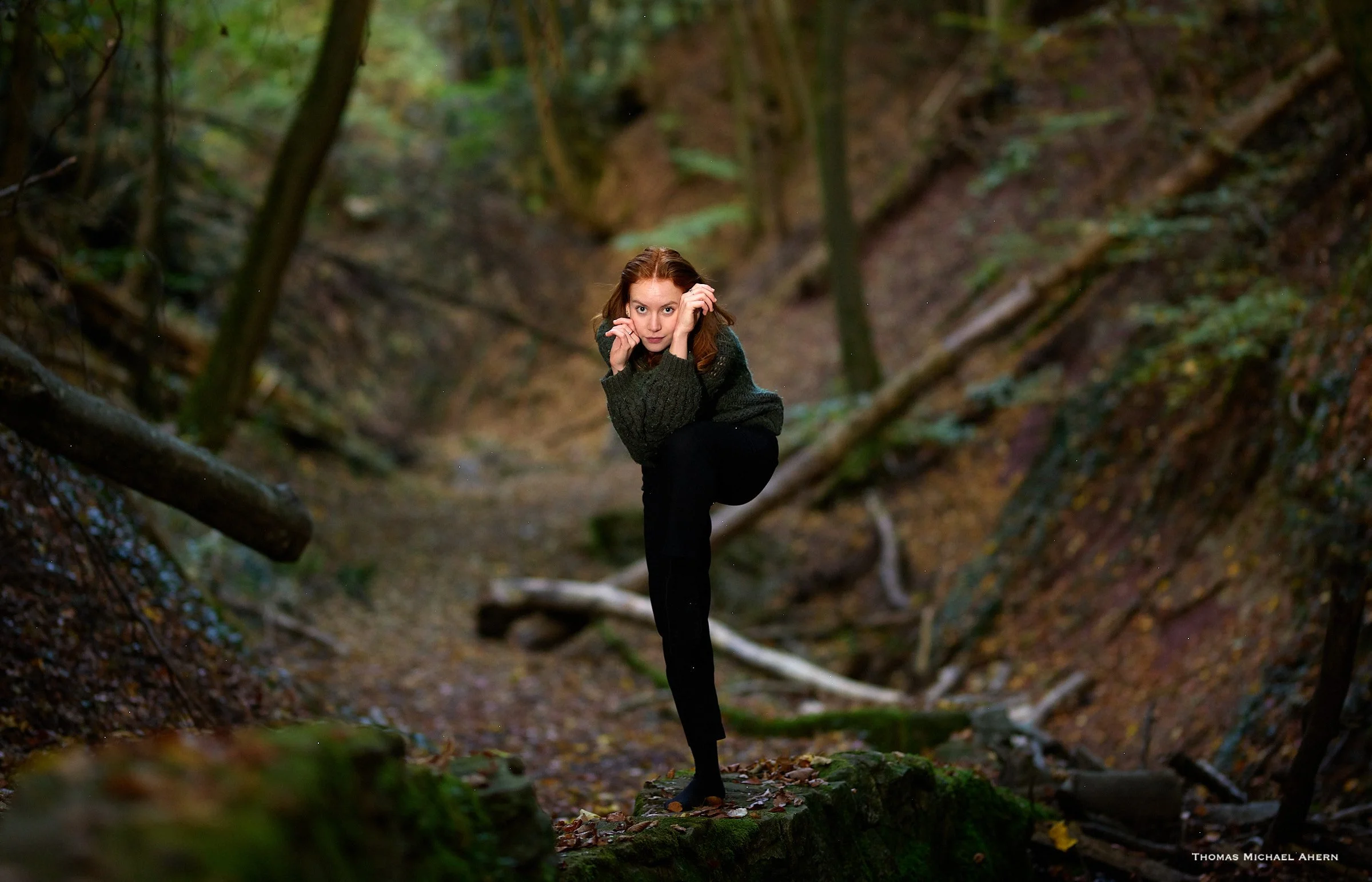 A woman with red hair and a dark sweater balancing on a fallen log in a dense forest with green foliage and tree trunks in the background.