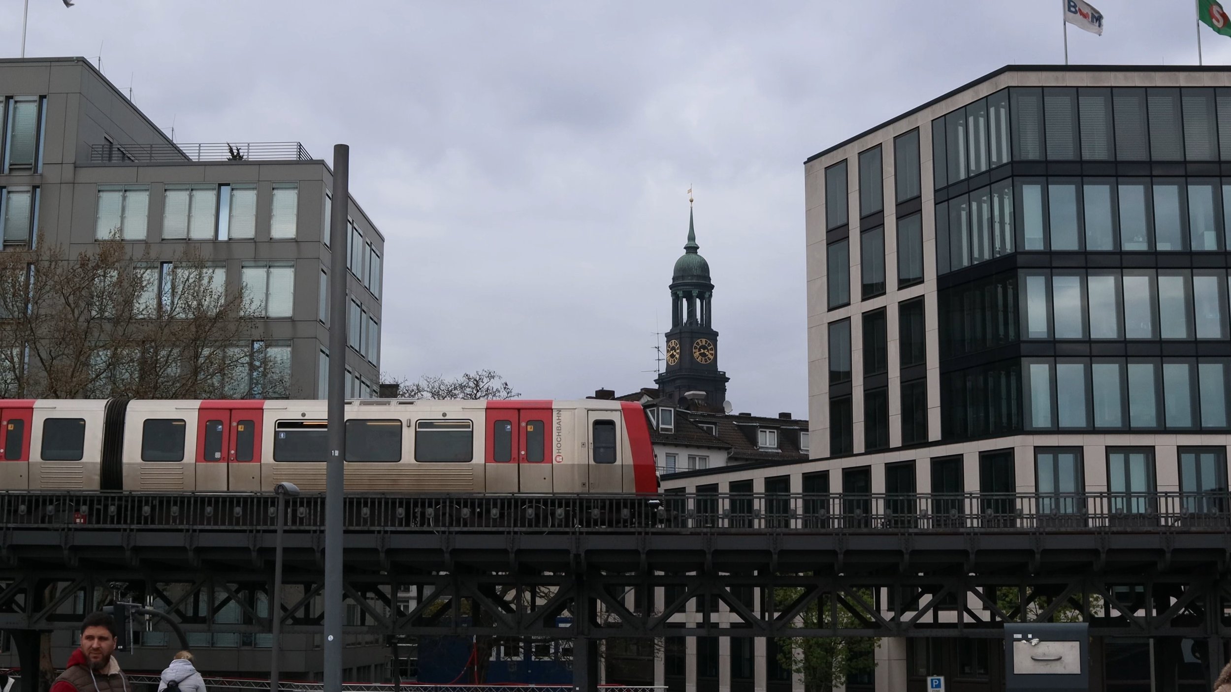 Urban scene with modern glass buildings, a historical clock tower, a train on an elevated track, and people walking on the street.
