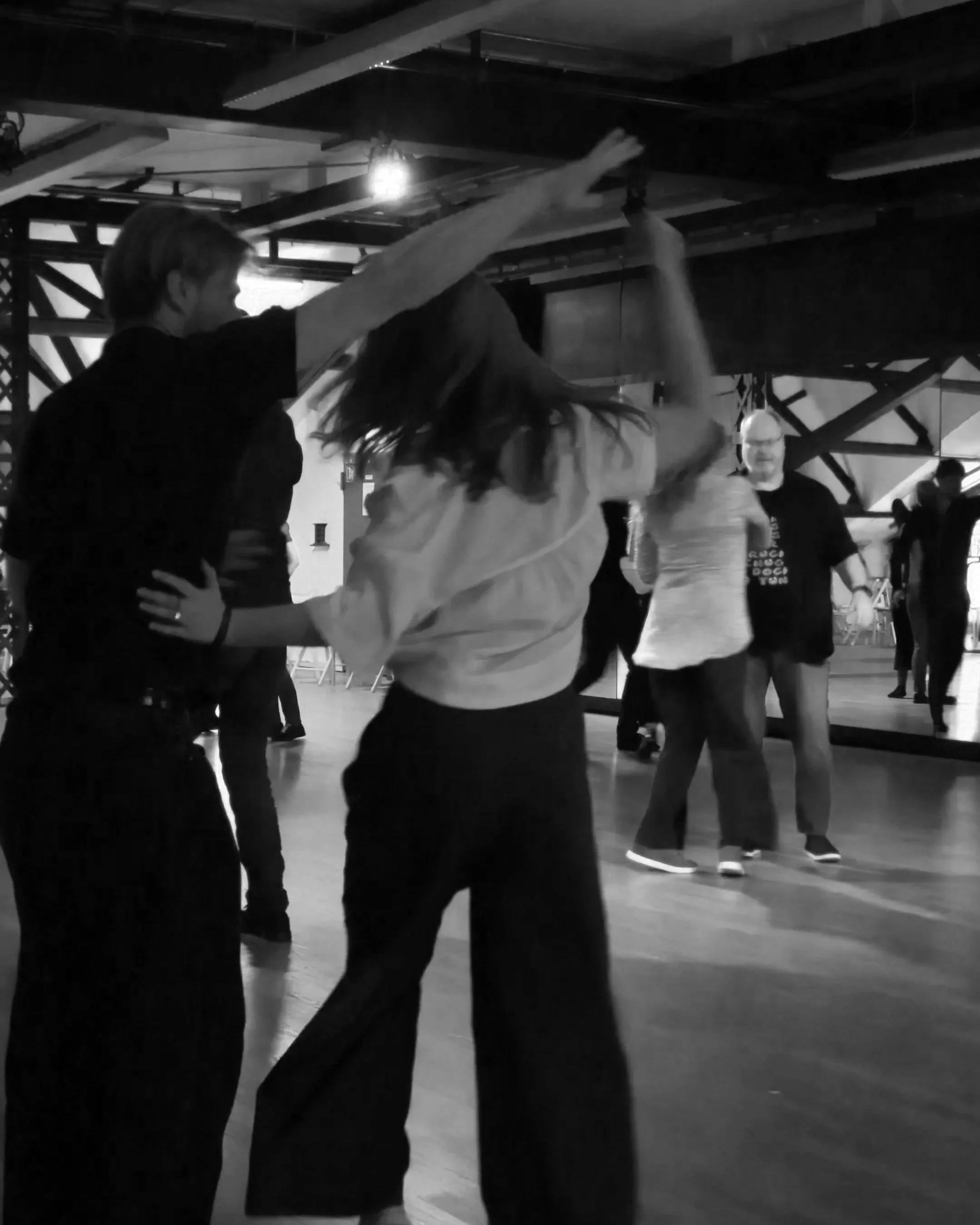 Two people dancing closely in a dimly lit indoor space, with other individuals in the background.