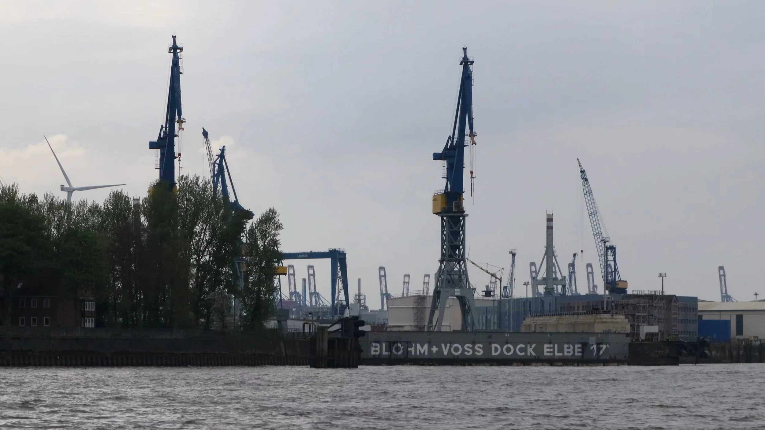 View of a harbor with several large blue and yellow crane structures and industrial buildings along the waterfront. The water in the foreground and trees in the left background.