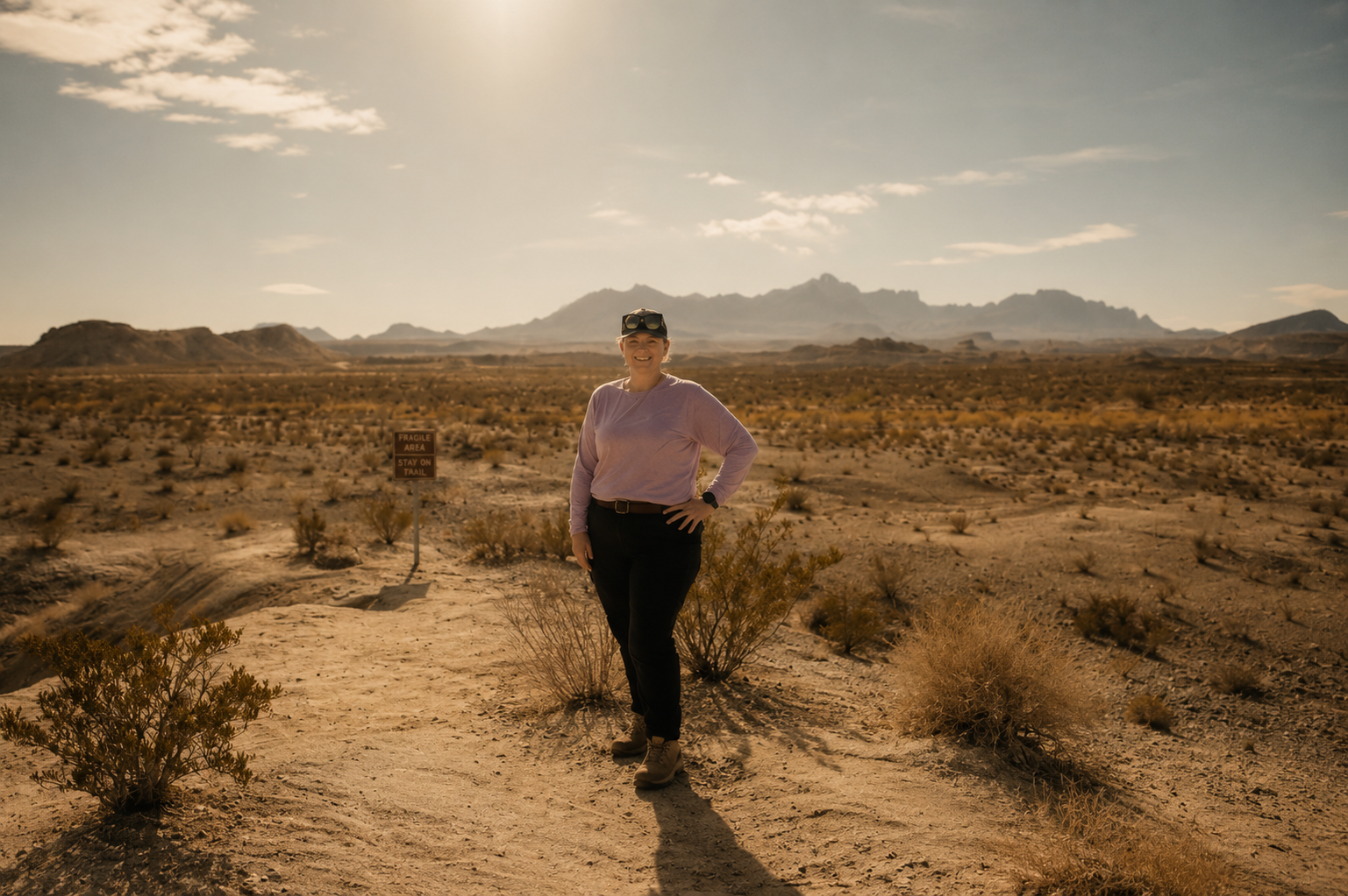 A woman stands in a desert landscape with mountains in the background, wearing a purple long-sleeve shirt, black pants, and sunglasses, smiling at the camera.