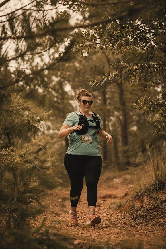 A woman running on a dirt trail through a wooded area wearing sunglasses, a teal shirt, black leggings, and brown running shoes, with a black hydration pack and a race bib numbered 196.