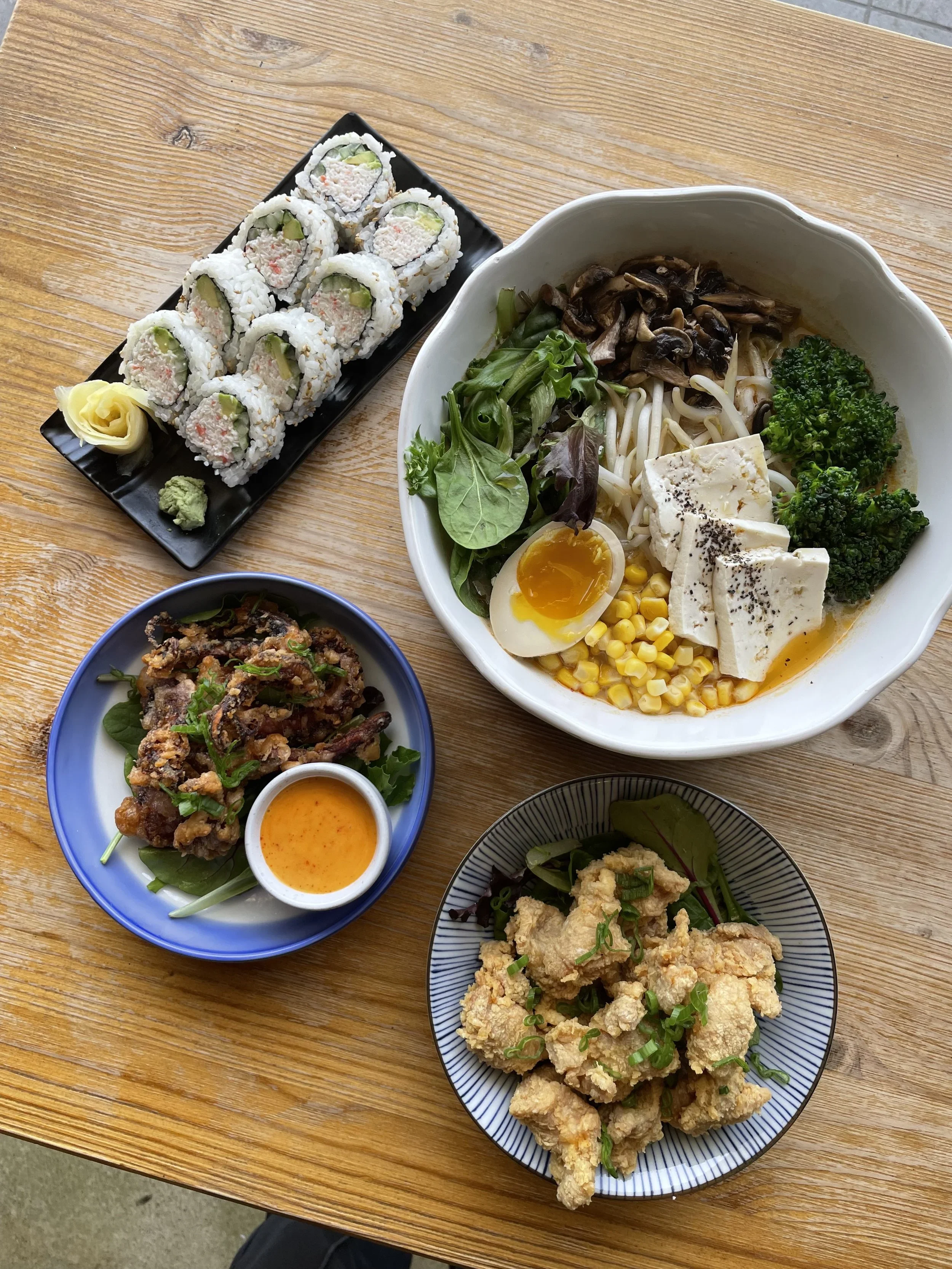 A variety of Asian dishes on a wooden table, including sushi rolls, salad with tofu, fried chicken, and fried fish, along with dipping sauces.