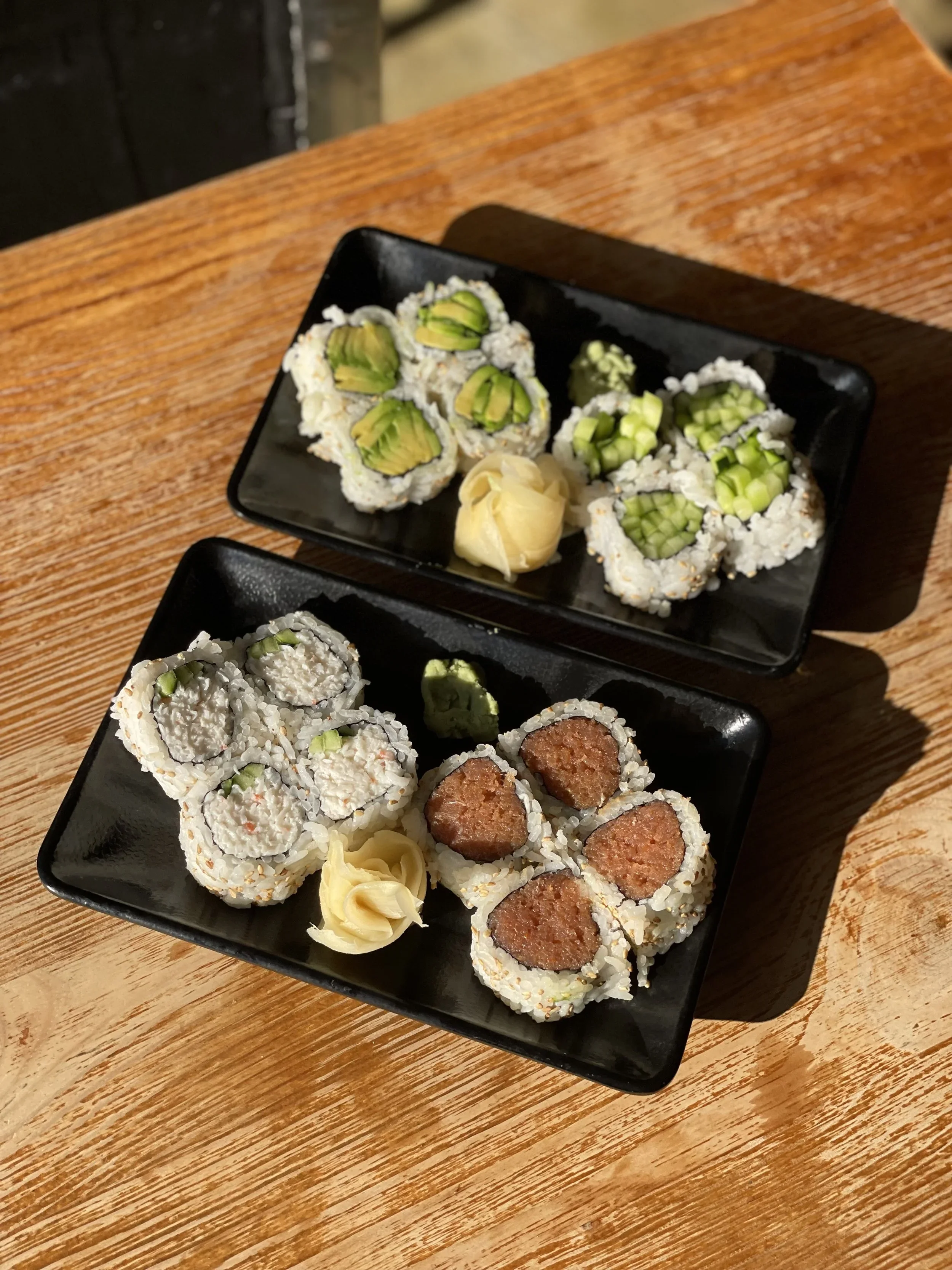 Two black rectangular plates containing sushi rolls with various fillings, garnished with pickled ginger and wasabi, placed on a wooden table.