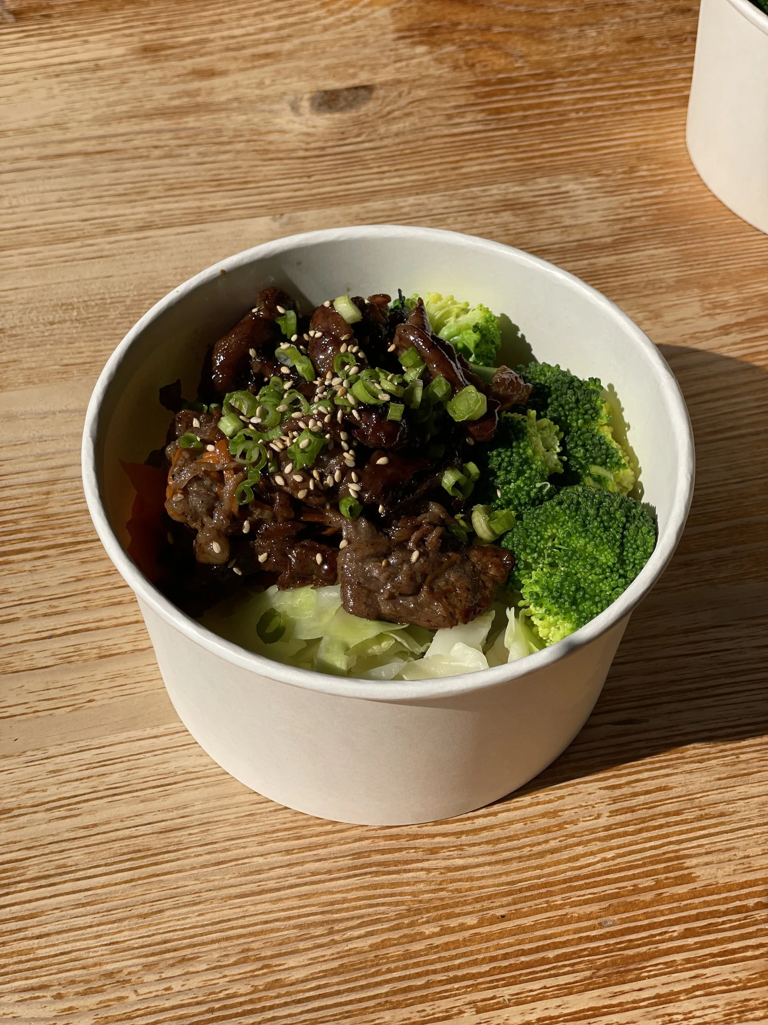 A white bowl containing beef, chopped green onions, sesame seeds, broccoli, and shredded lettuce on a wooden table.