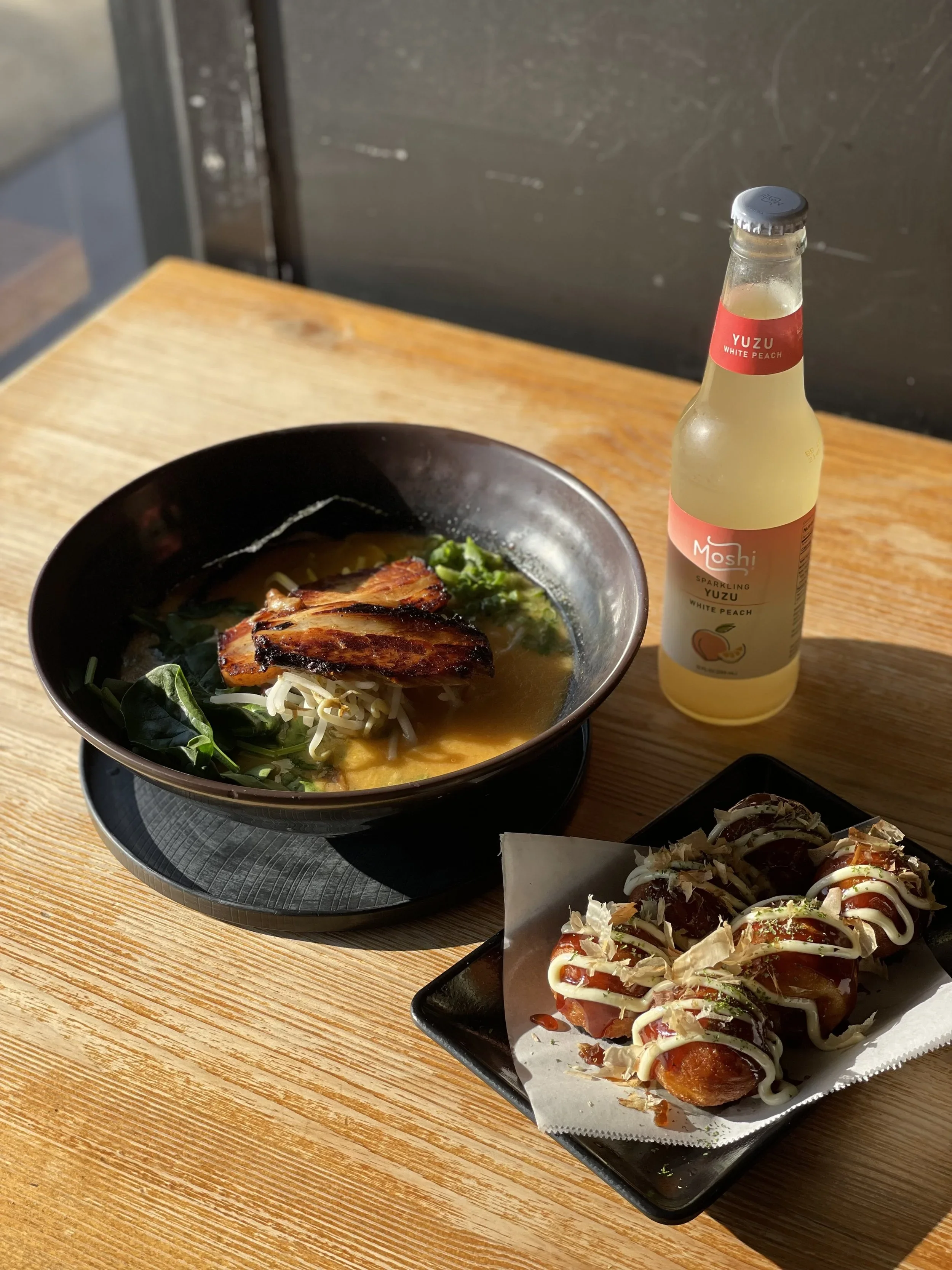 A bowl of ramen noodles with grilled chicken, green vegetables, and broth, alongside a bottle of sparkling yuzu white peach drink, and a plate of sushi rolls with sauce and toppings on a wooden table.