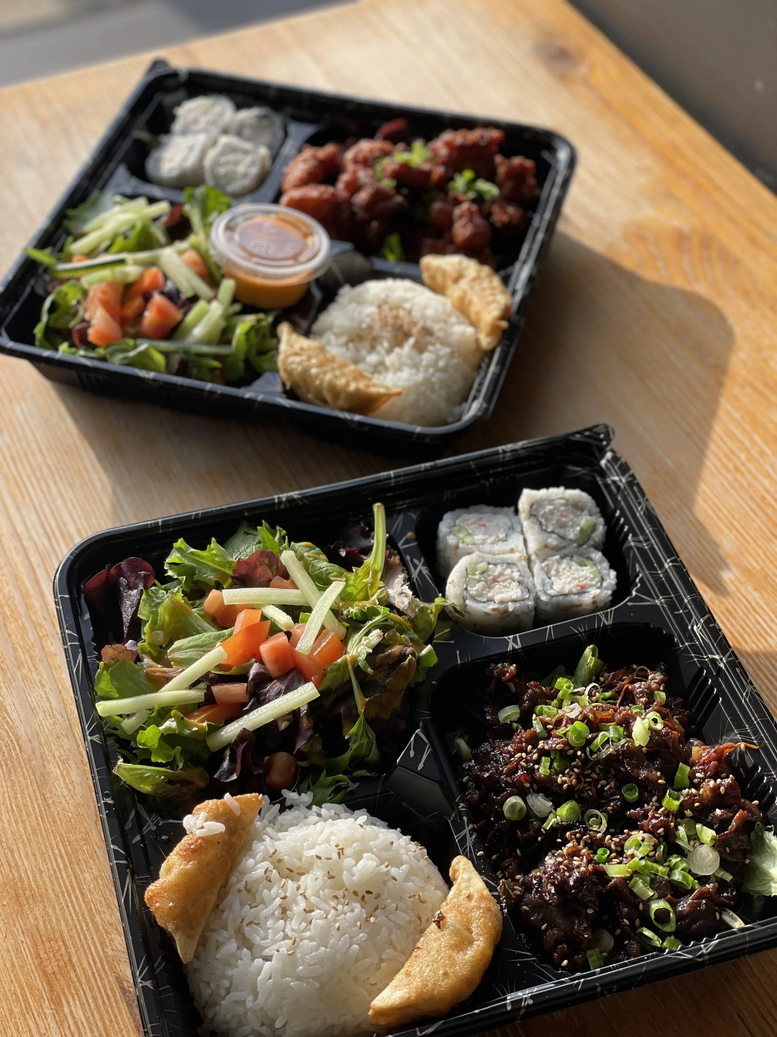 A black plastic bento box divided into sections contains a variety of Asian dishes. The sections include rice, spring rolls, a mixed vegetable salad, sushi rolls, and a portion of stir-fried beef with green onions garnished with sesame seeds.