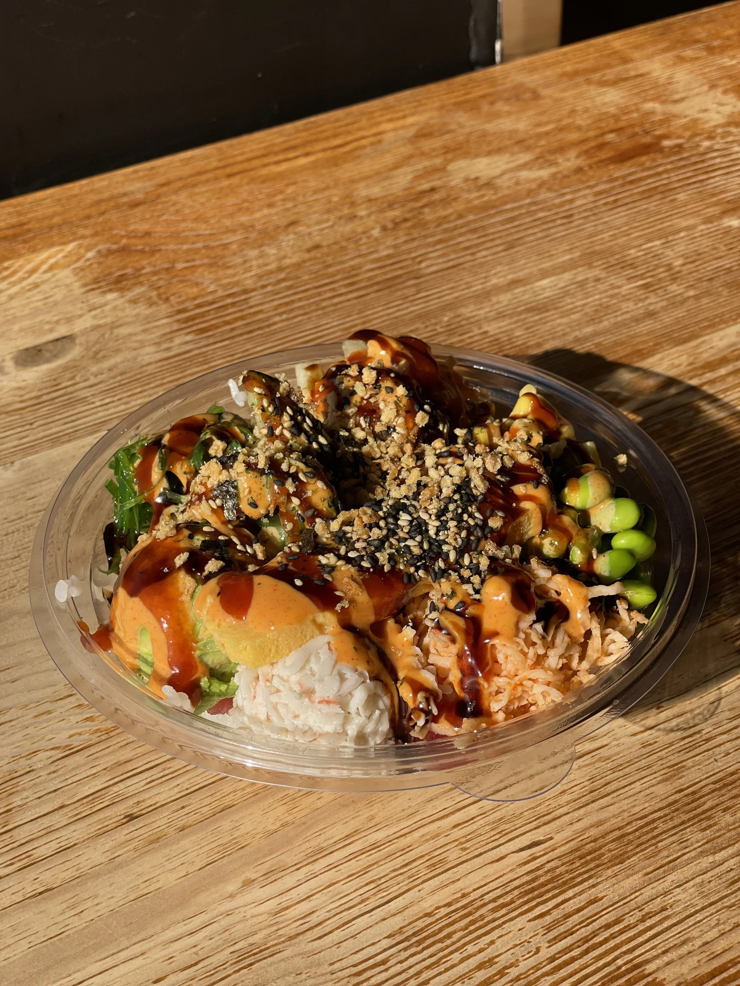 Mixed sushi salad in a clear plastic bowl with various sushi ingredients, topped with sauces and sesame seeds, placed on a wooden table.