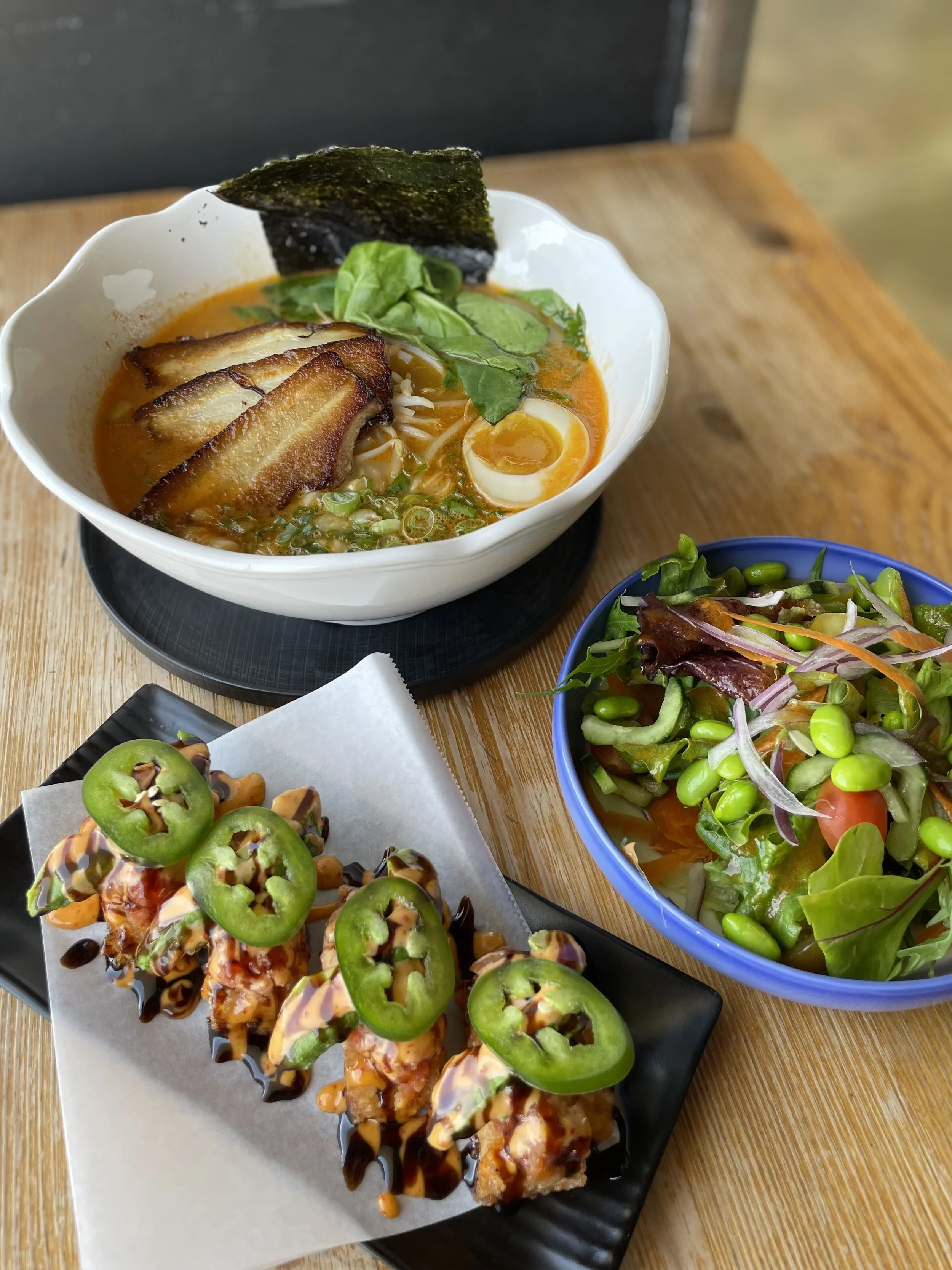 A bowl of spicy ramen topped with sliced pork, a halved soft-boiled egg, seaweed, and greens, served on a black tray, alongside a colorful salad with greens, celery, and cherry tomatoes, on a wooden table.