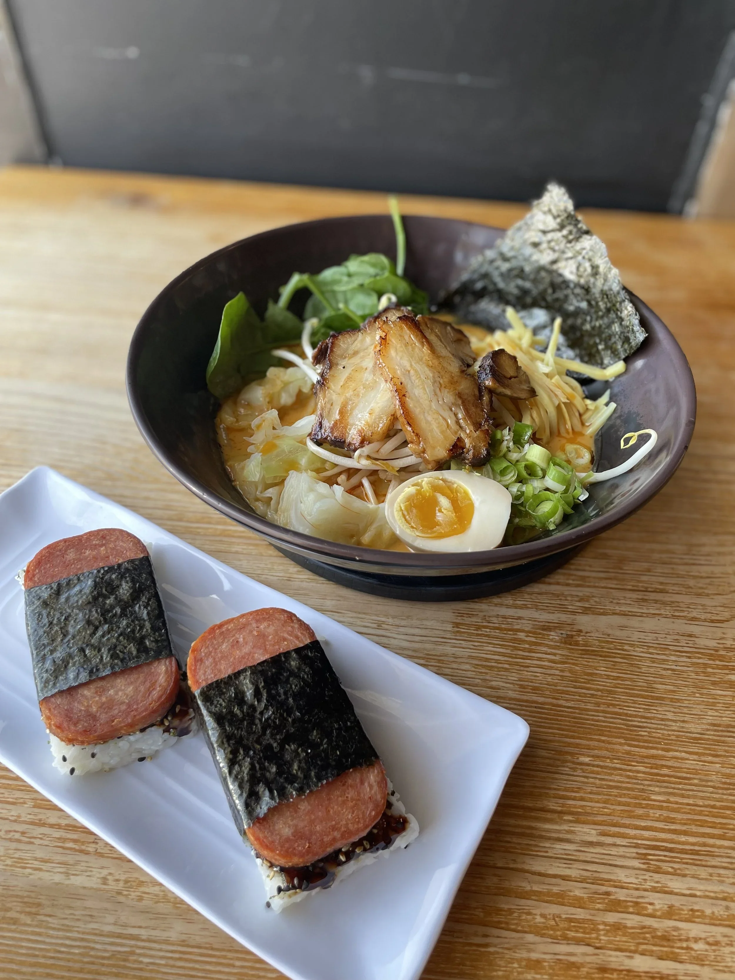 A bowl of ramen with boiled egg, vegetables, pork belly, and seaweed, alongside sushi rolls with rice, seaweed, and sliced meat.