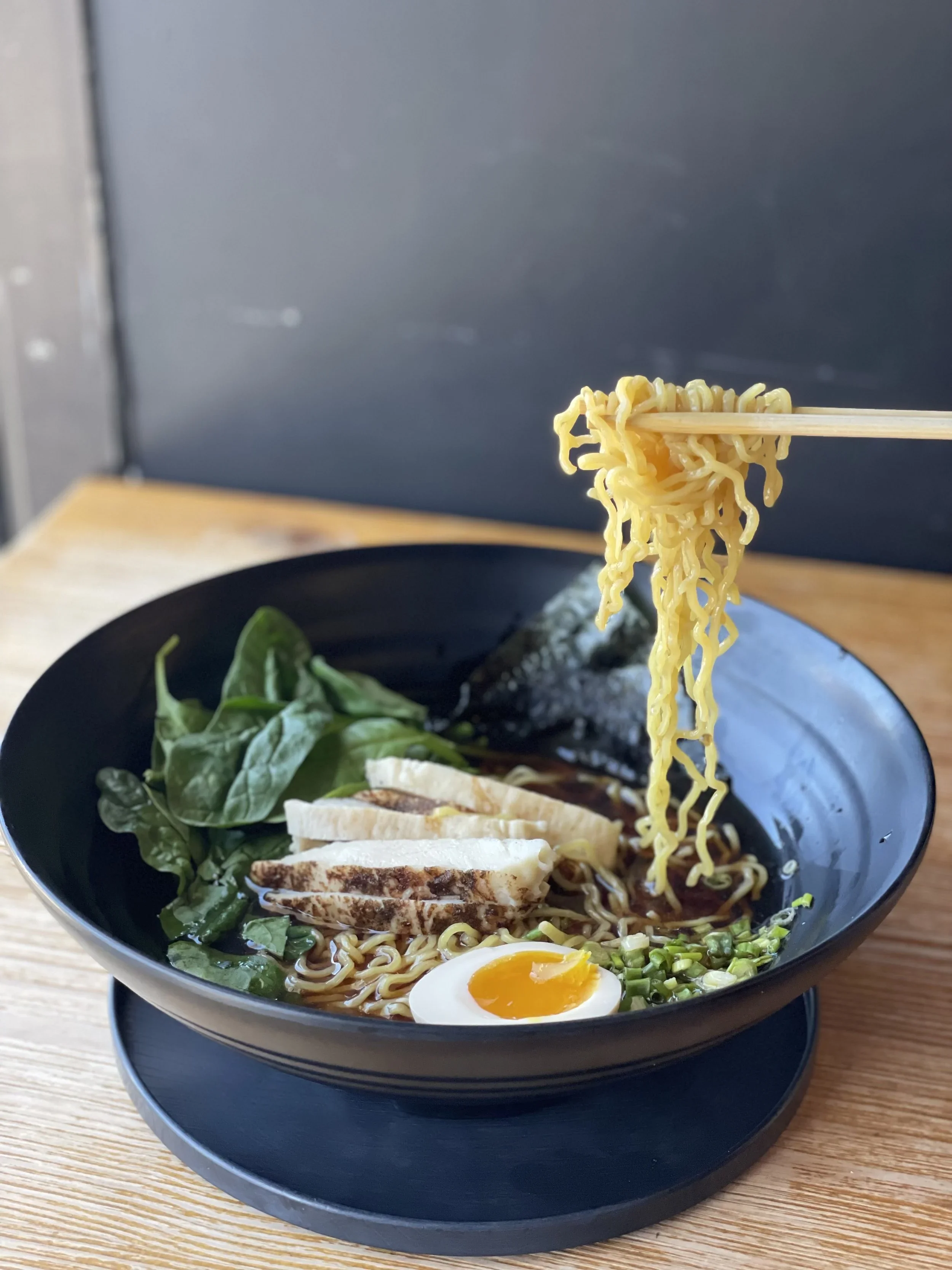Bowl of ramen noodles with sliced grilled chicken, half a soft-boiled egg, spinach, chopped scallions, and seaweed on a wooden table.