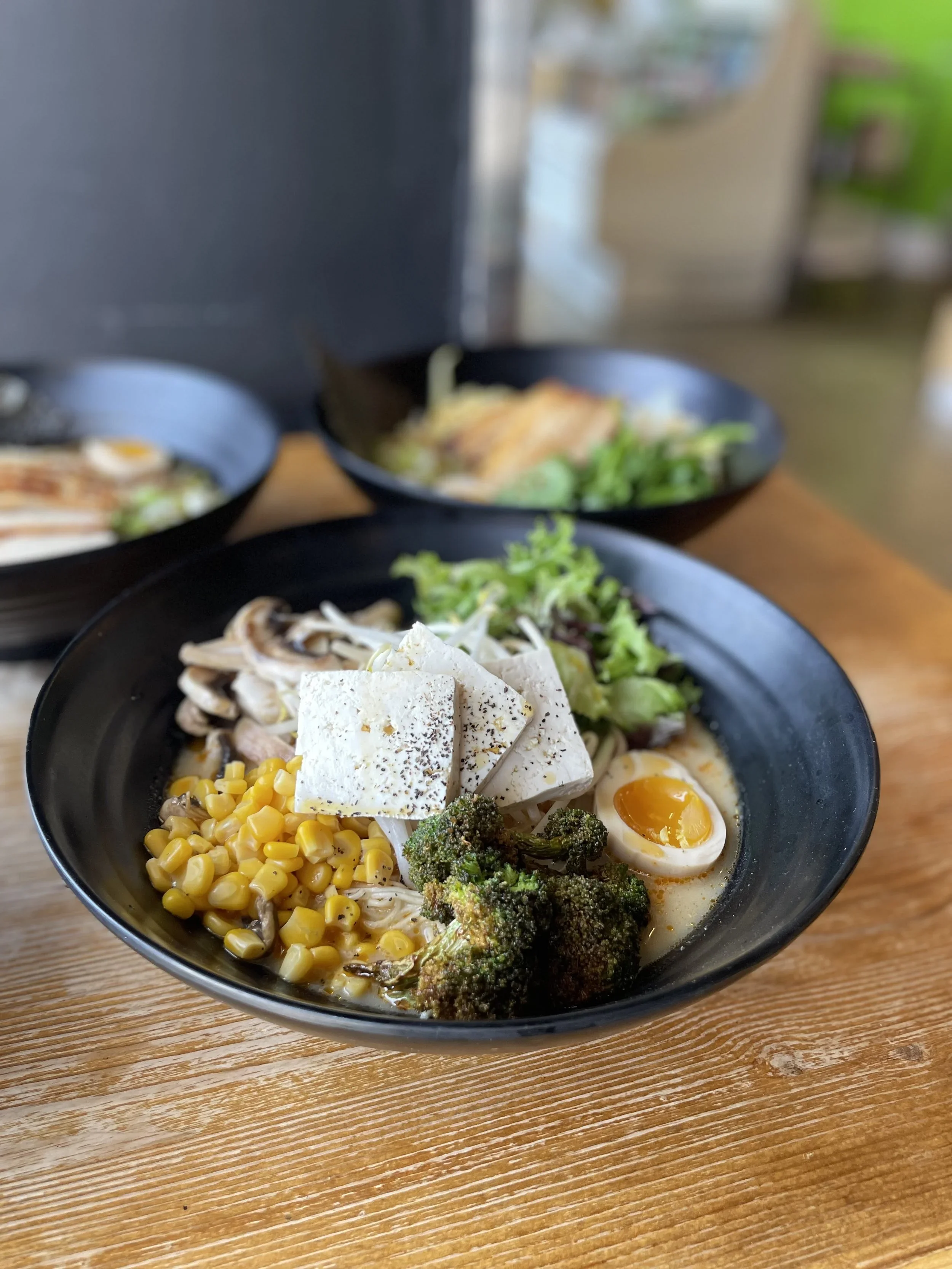 A black bowl of ramen with corn, broccoli, sliced mushrooms, shredded lettuce, tofu, and soft-boiled eggs on a wooden table.