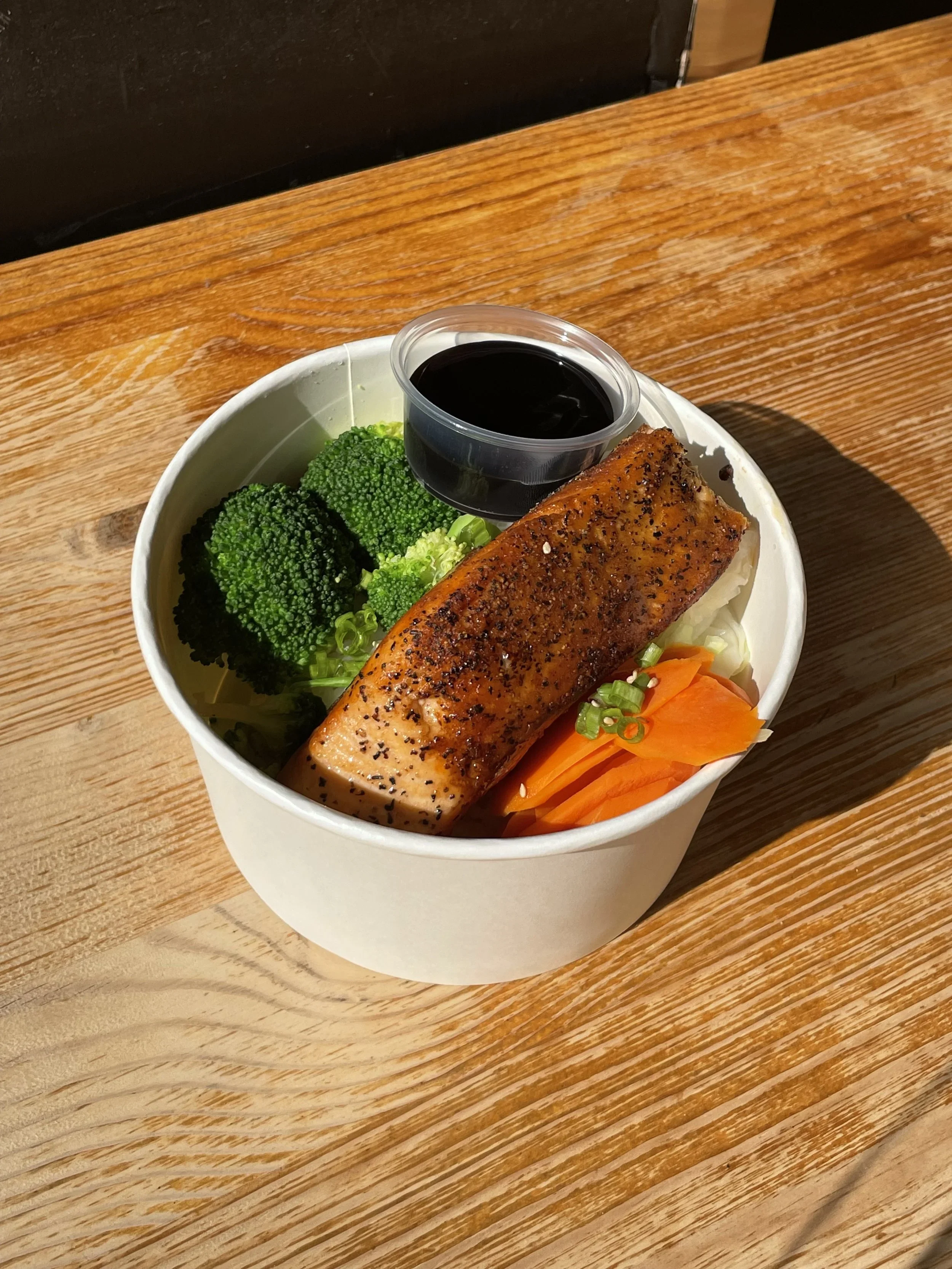 Bowl of salmon, broccoli, carrots, and sauce on a wooden table.