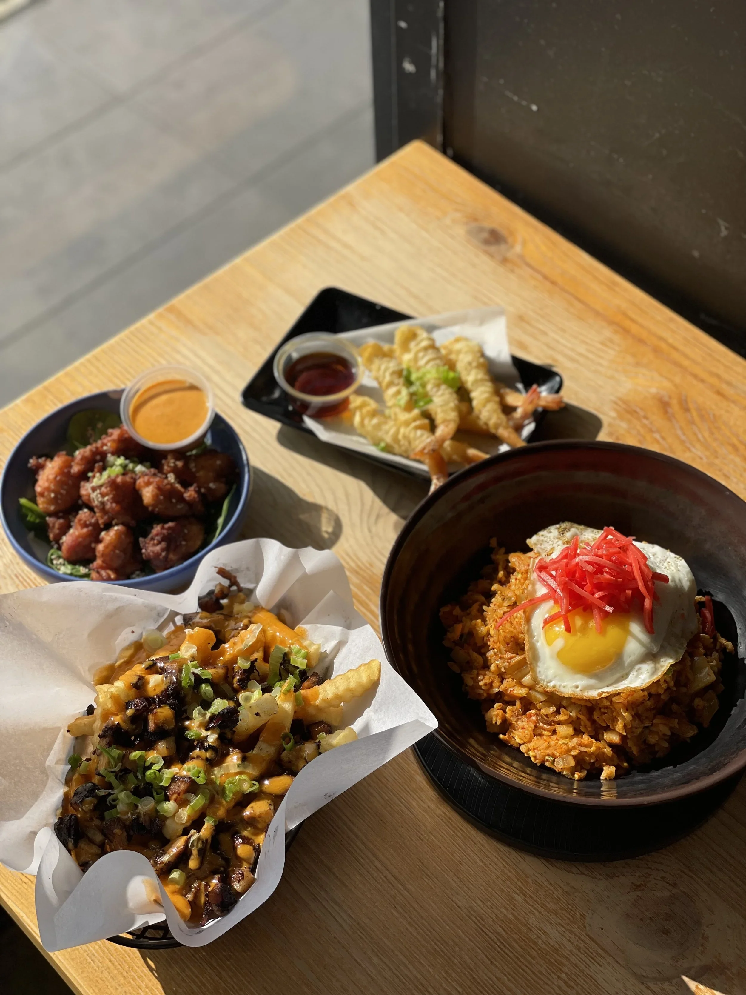 Assorted Asian dishes on a wooden table, including fried shrimp with dipping sauces, fried chicken pieces, a rice bowl with a fried egg and toppings, and a taco topped with cheese and green onions.