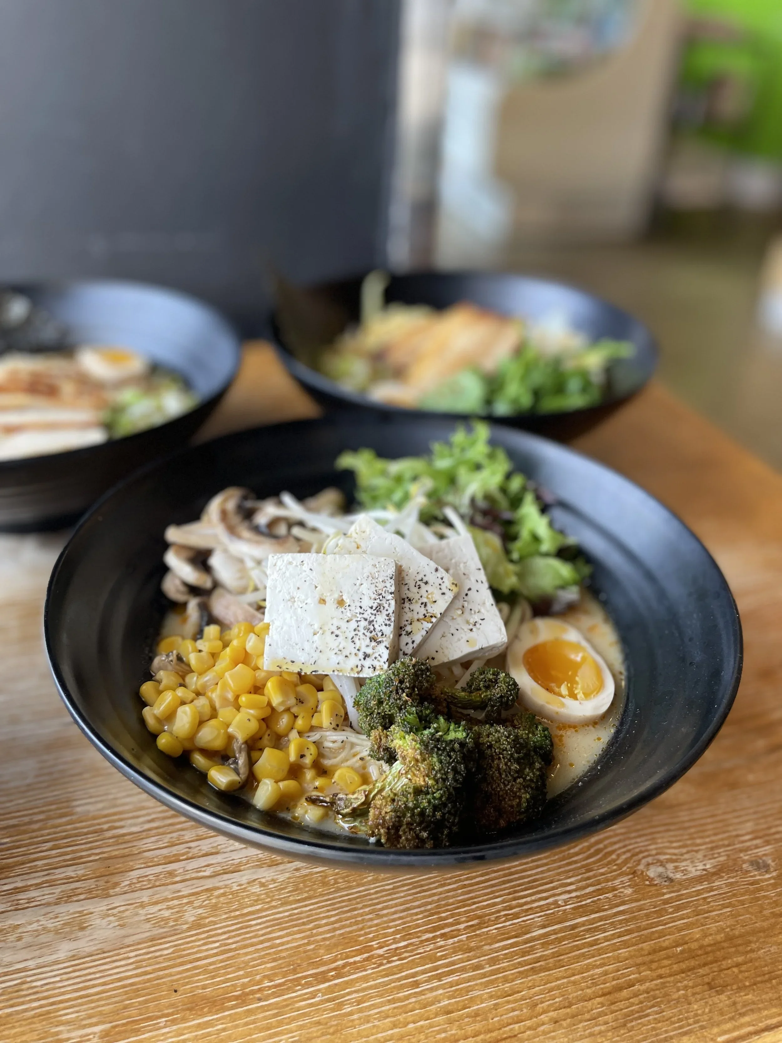 Bowl of ramen with corn, tofu, broccoli, boiled egg, and shredded chicken on a wooden table.