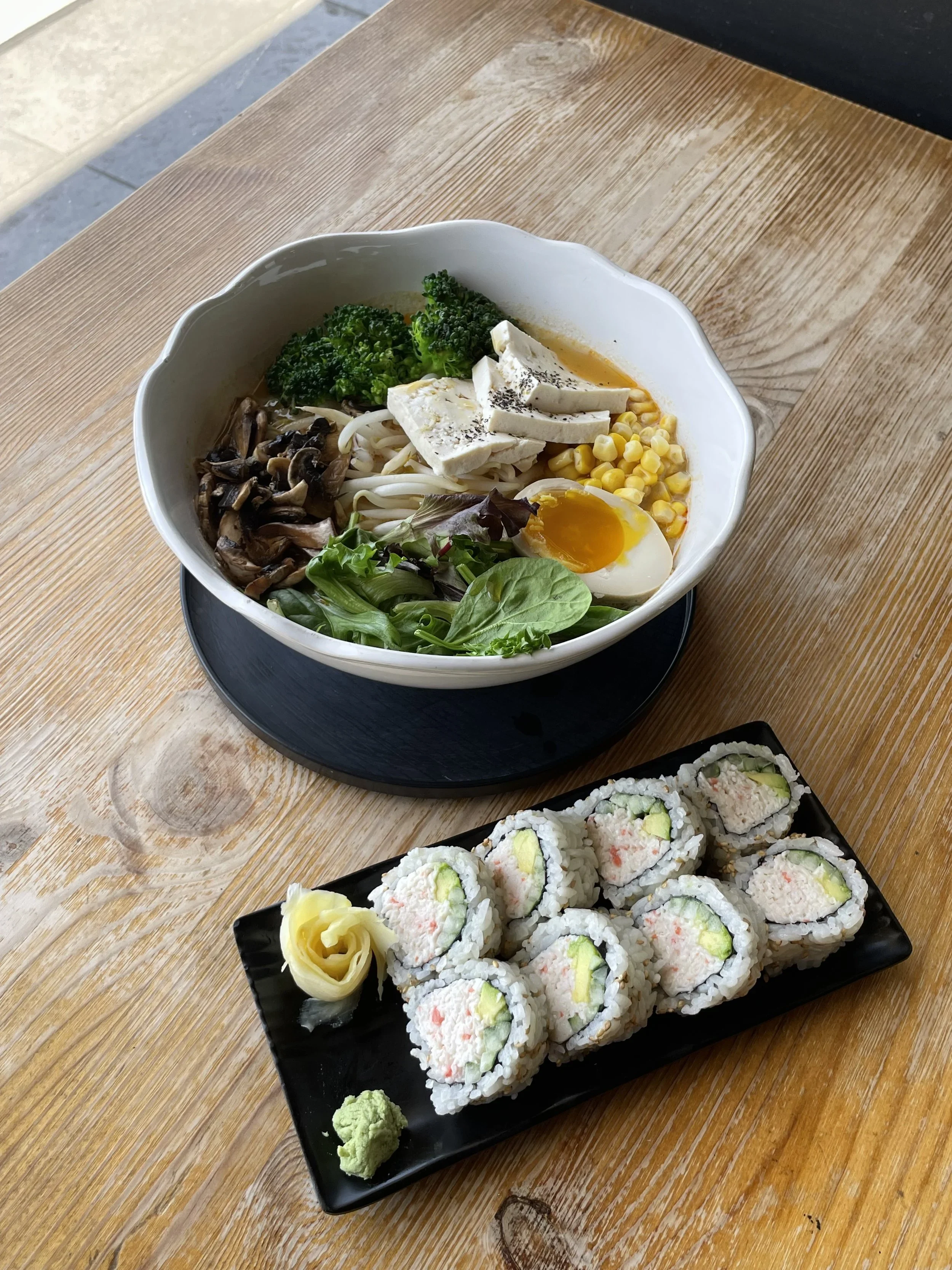 A bowl of ramen with broccoli, tofu, corn, mushrooms, bean sprouts, and half a boiled egg, served on a wooden table. A plate of sushi rolls with avocado, imitation crab, and cucumber, along with wasabi and pickled ginger.