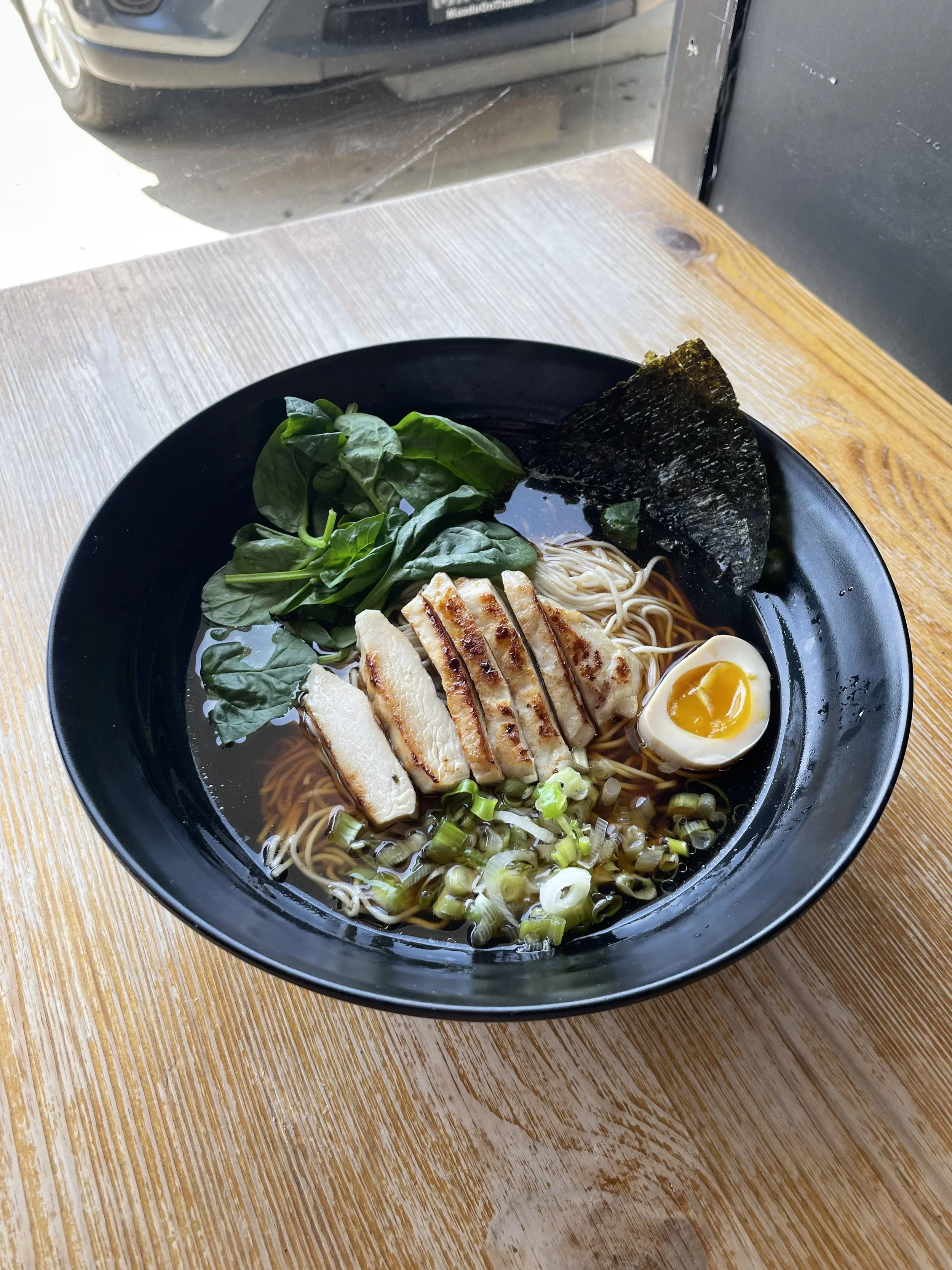 A bowl of ramen with sliced grilled chicken, half a soft-boiled egg, seaweed, green leafy vegetables, chopped green onions, and noodles in broth on a wooden table.