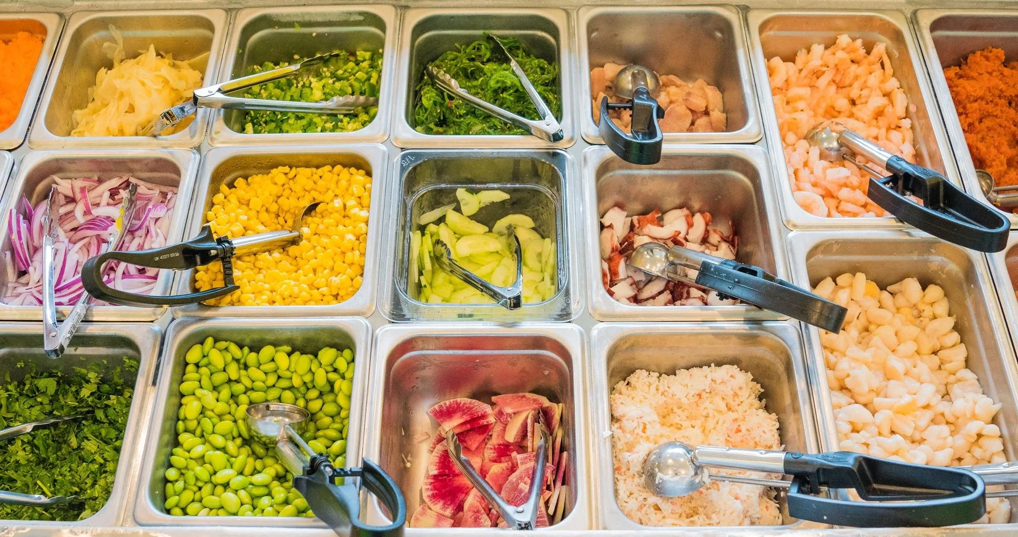 Assorted fresh vegetables in a self-serve salad bar, including sliced onions, chopped green onions, seaweed, chicken, shrimp, corn, sliced cucumbers, edamame, cilantro, sliced watermelon, and shredded cheese, with tongs and serving utensils.