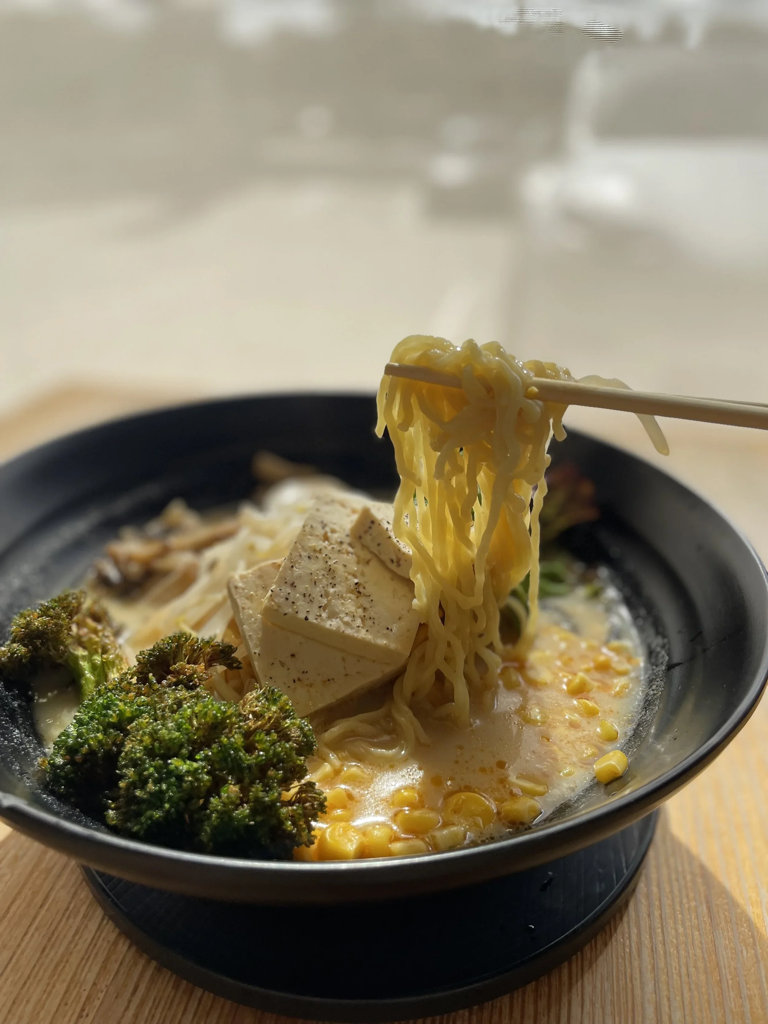 Close-up of a bowl of ramen noodles with tofu, broccoli, and corn, with chopsticks lifting noodles. The bowl is on a black tray on a wooden table.