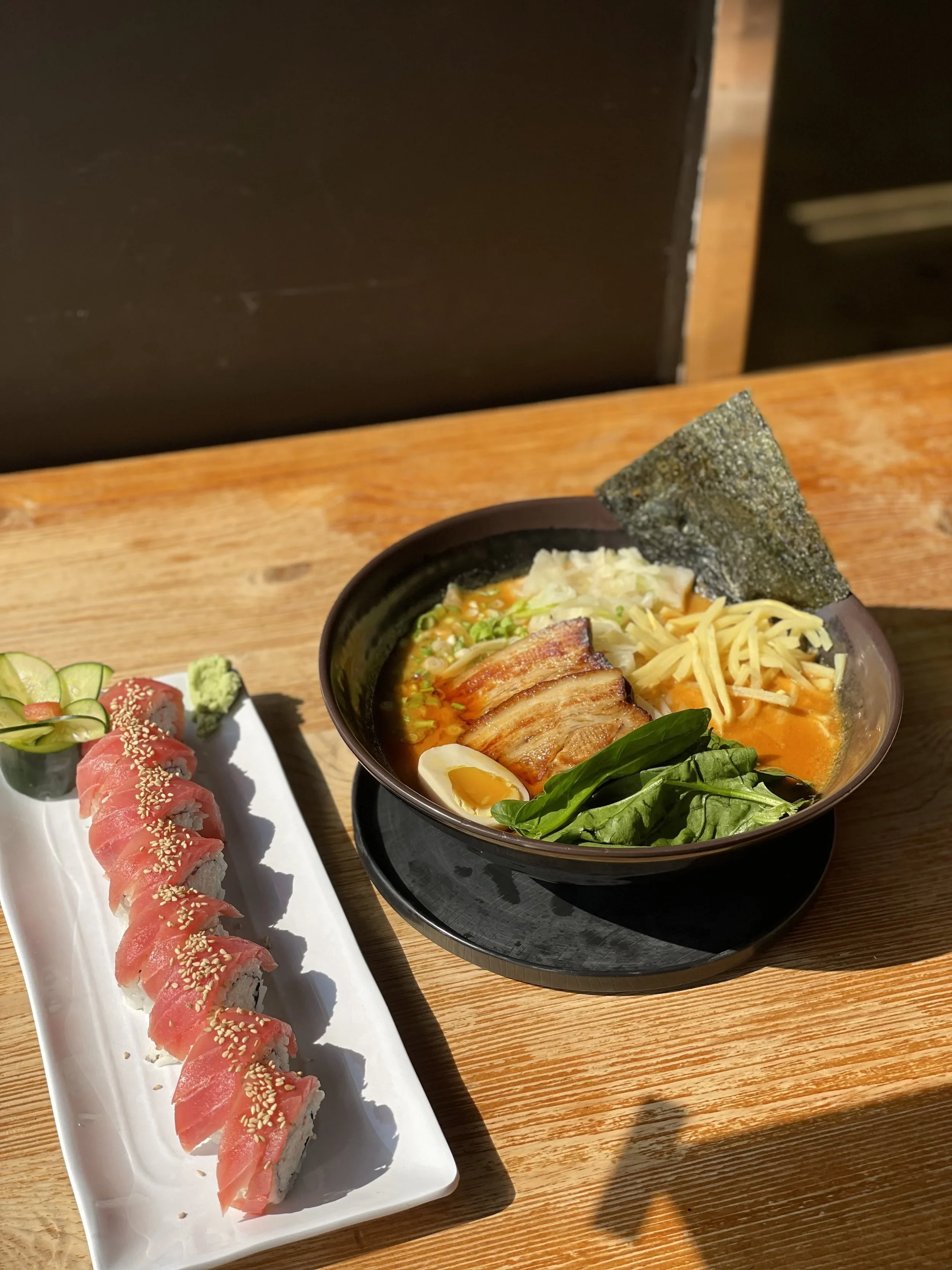 A black bowl of ramen with pork, green onions, seaweed, spinach, and half a soft egg, with a white rectangular plate of sushi rolls topped with tuna, sesame seeds, sliced cucumbers, and wasabi on a wooden table.