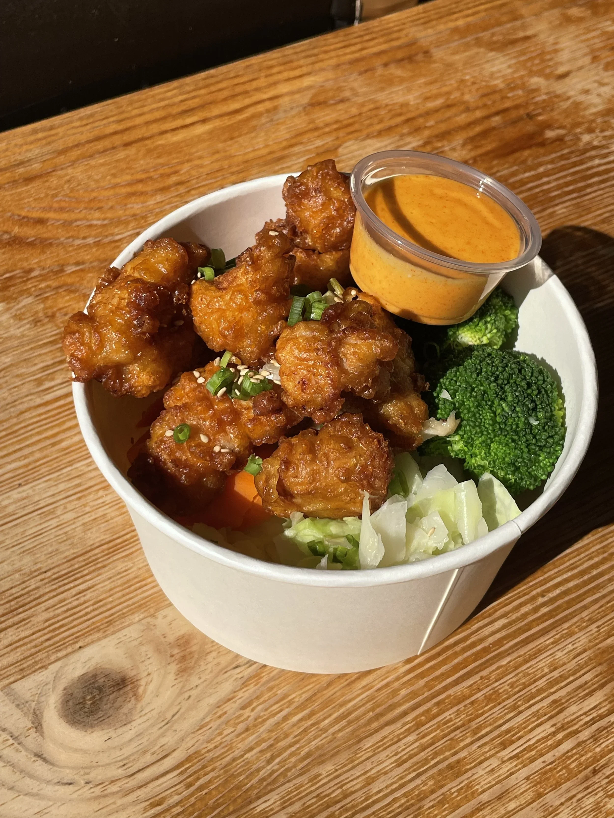 Fried chicken bites garnished with green onions, served with broccoli, a side of chopped cabbage, and a cup of orange-colored dipping sauce in a white bowl on a wooden table.