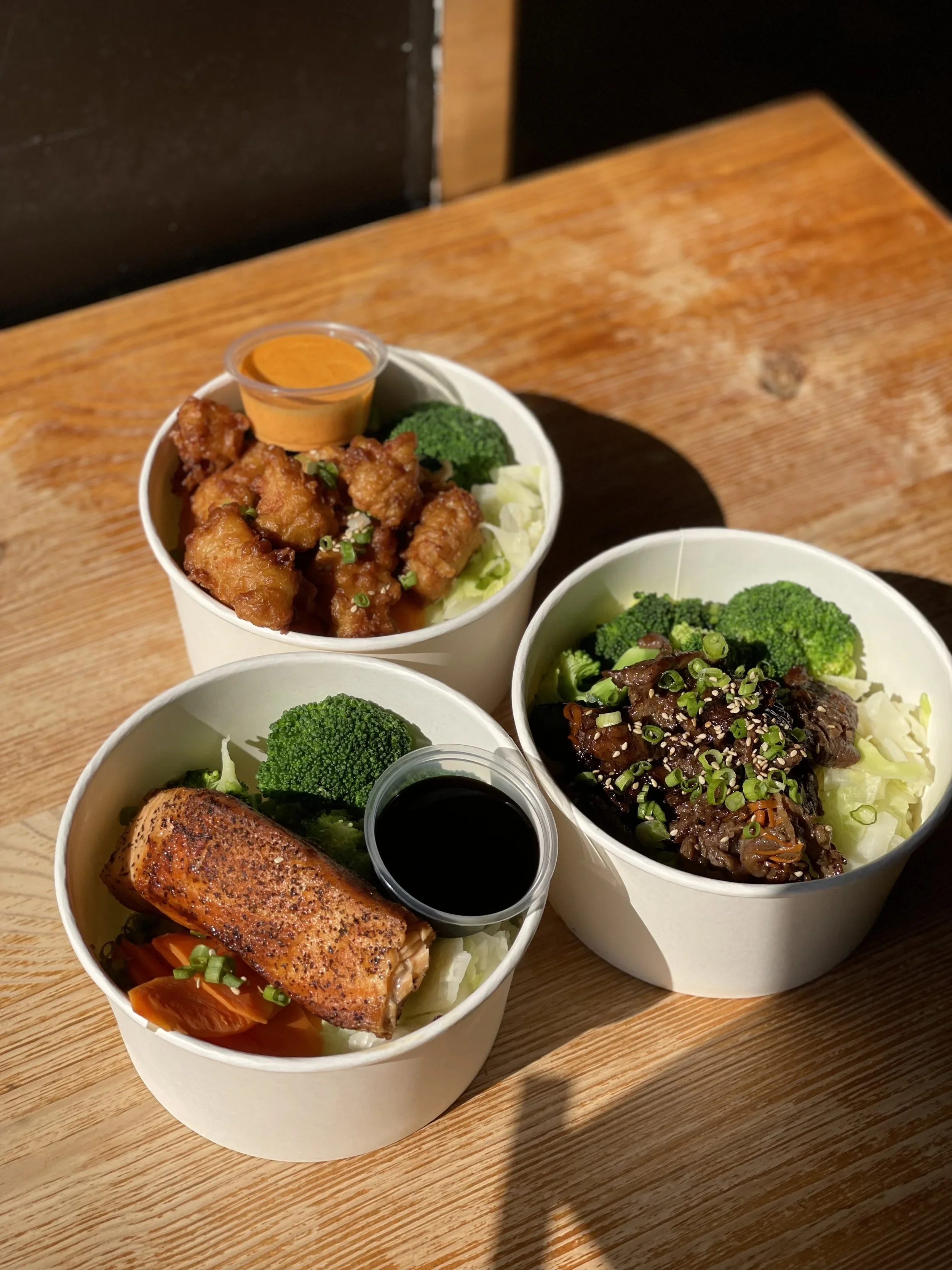Three bowls of Asian cuisine on a wooden table with broccoli, salmon, chicken, and rice, accompanied by sauces.