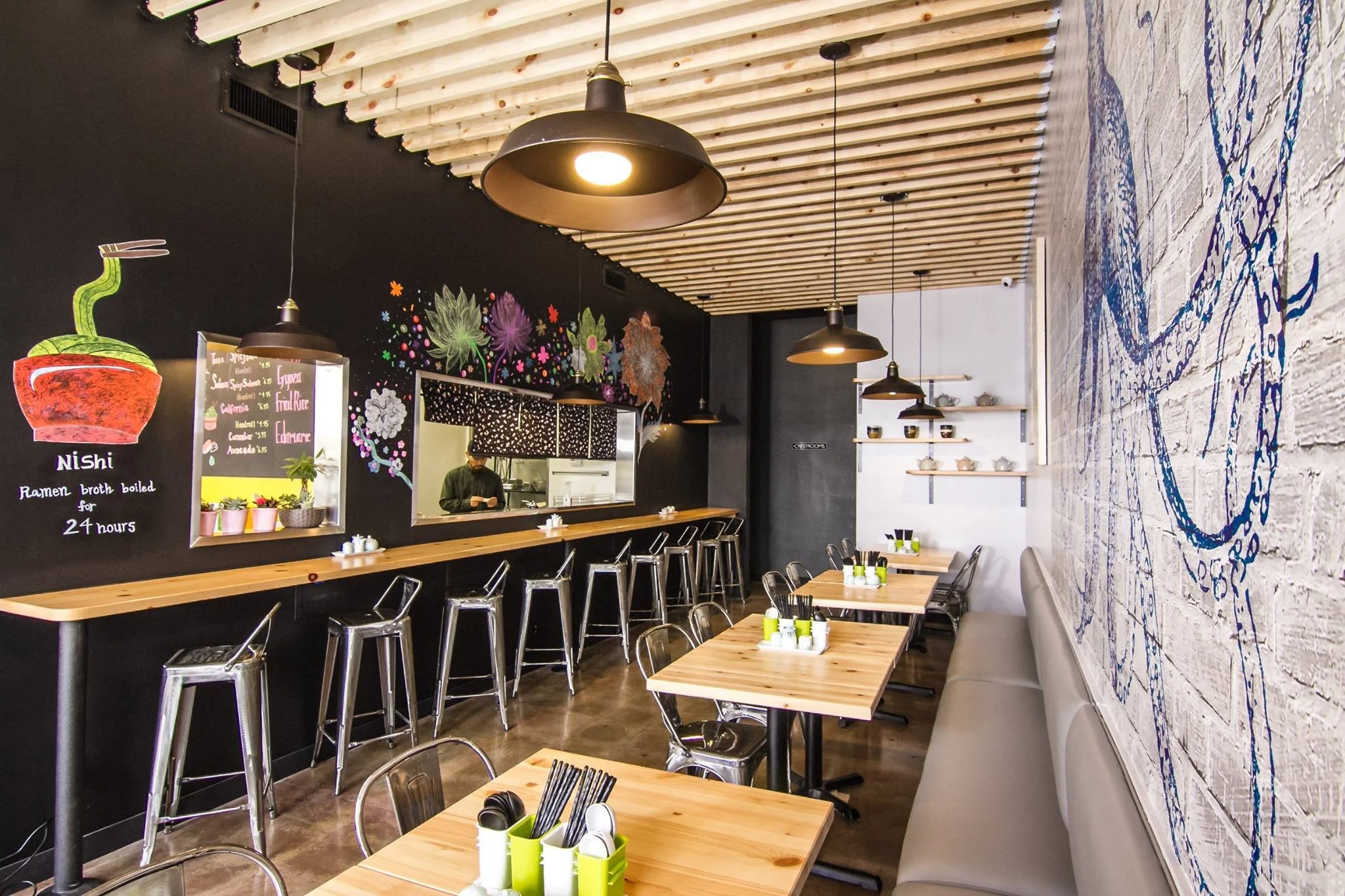 Interior of a modern restaurant with wooden tables, metal chairs, and playful wall art including a chalkboard menu and colorful floral drawings on the black wall. Pendant lights hang from a ceiling with exposed wooden beams.