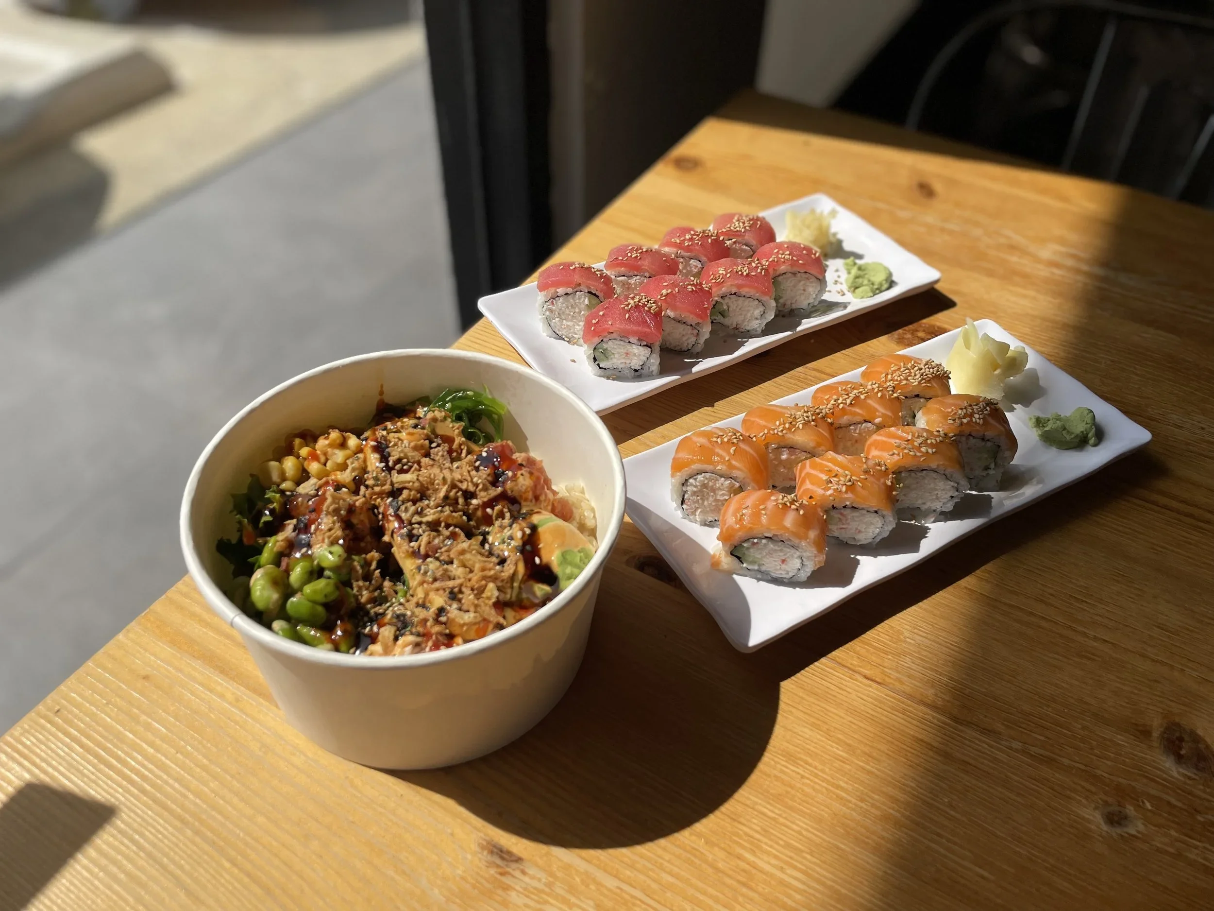 A wooden table with two rectangular dishes of sushi rolls, a bowl of Asian salad, and condiments of wasabi and ginger.