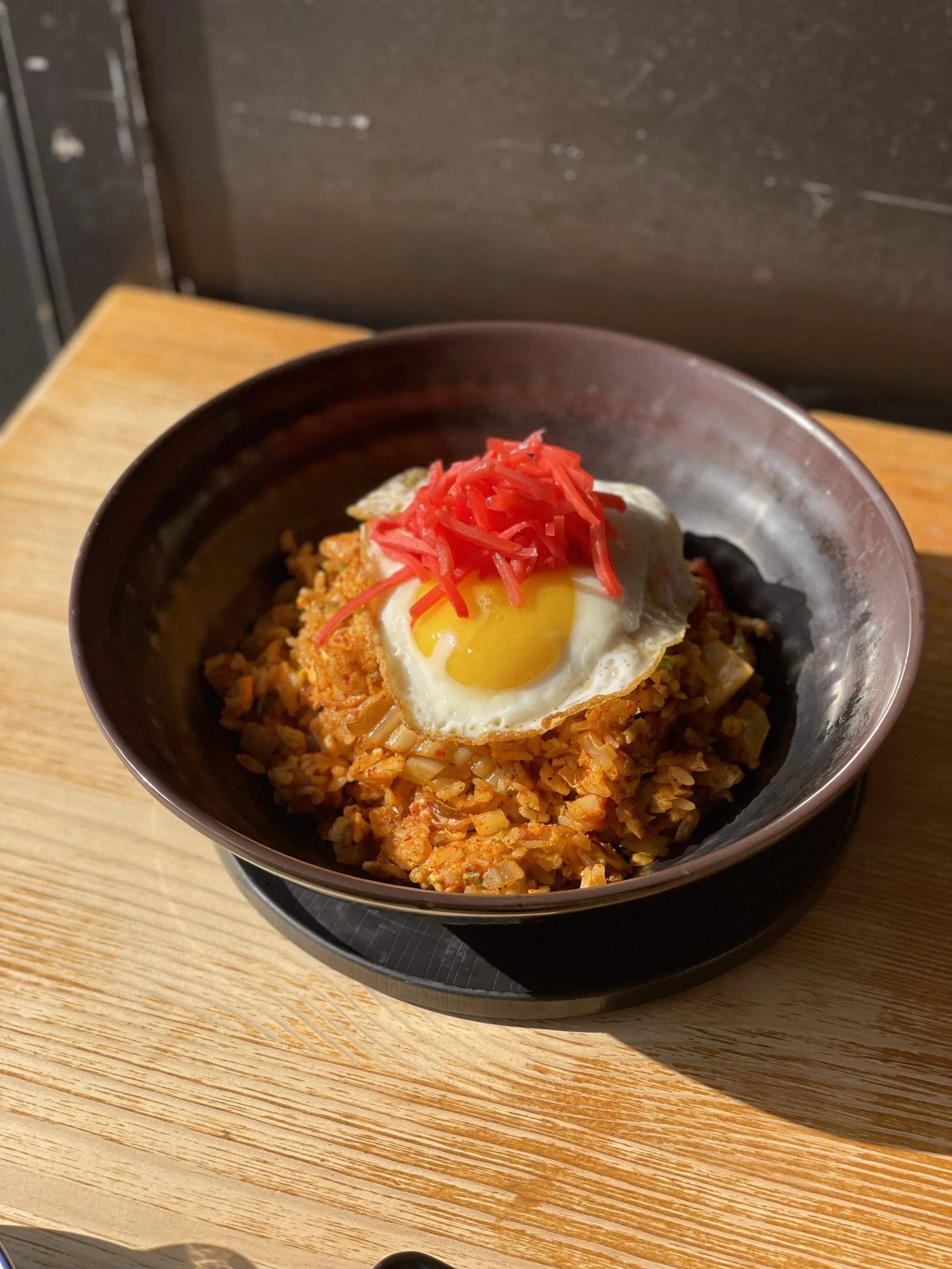 A bowl of fried rice topped with a sunny-side-up egg and shredded red pickled ginger, placed on a black tray on a wooden table.