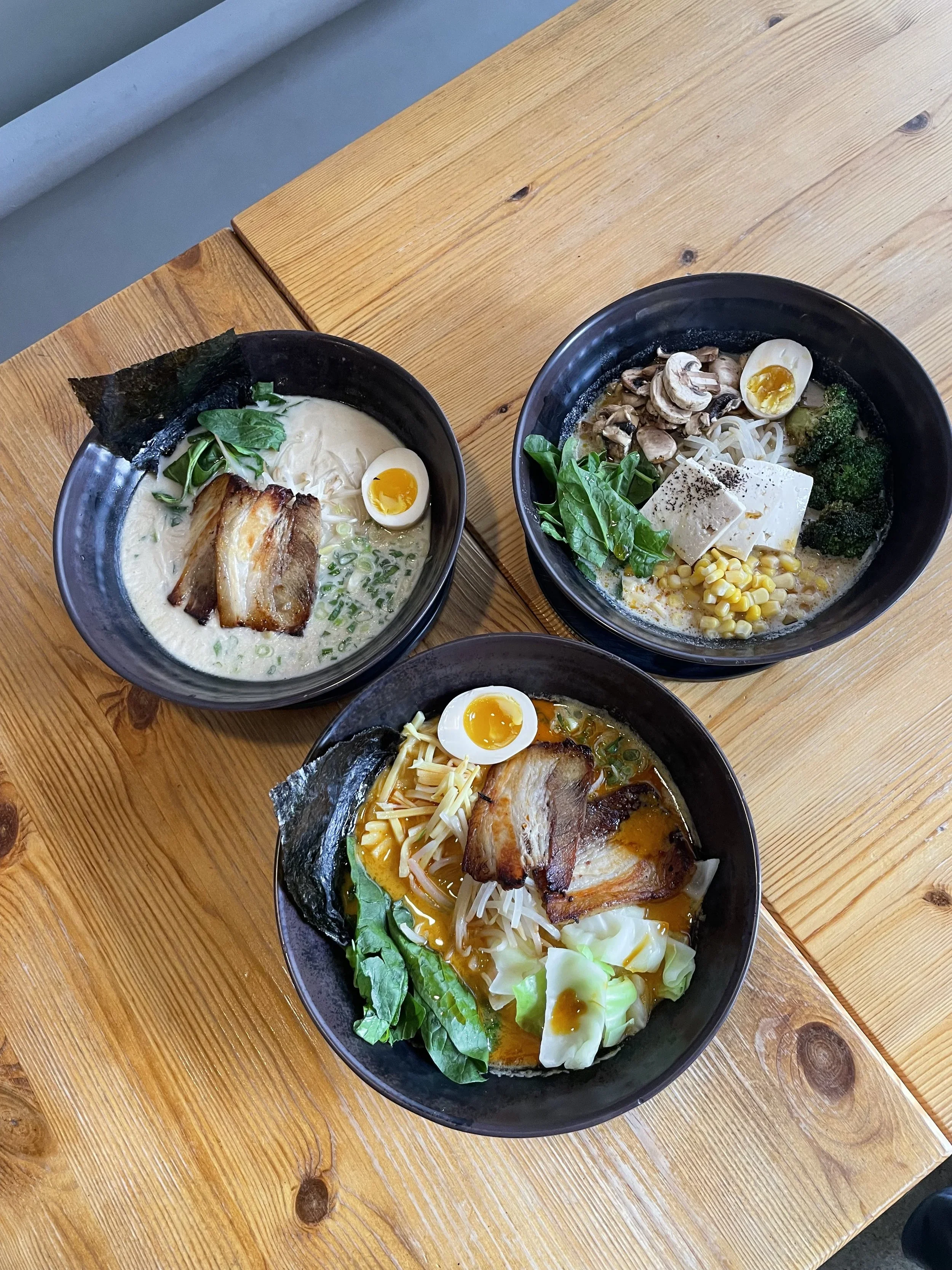 Three bowls of ramen with various toppings on a wooden table.