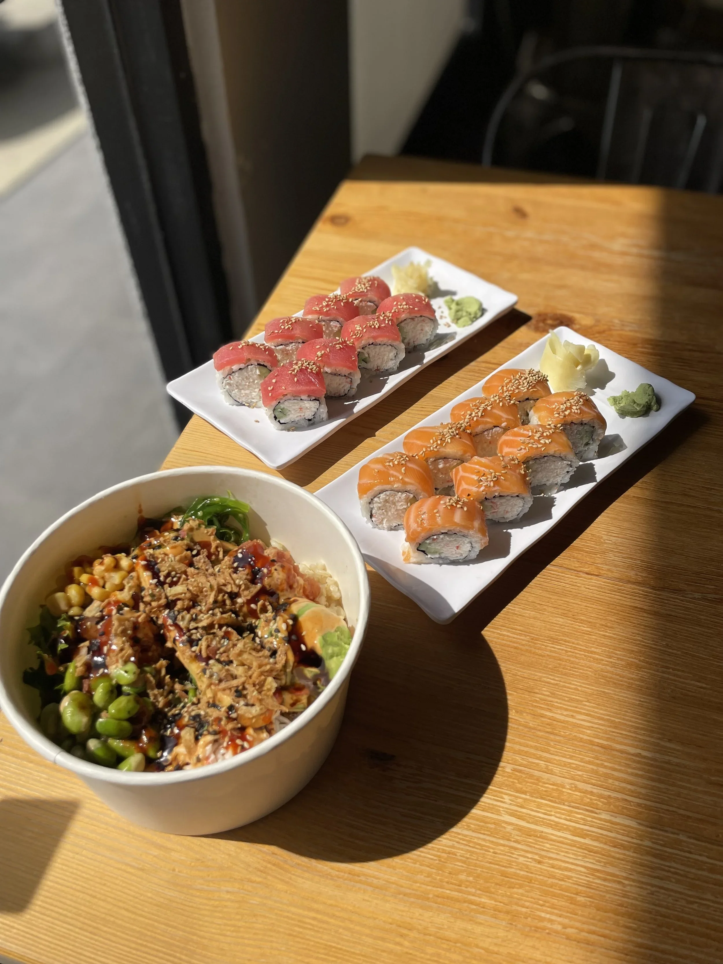 Assorted sushi rolls on white plates with wasabi, ginger, and a bowl of poke or rice bowl with vegetables in the foreground.