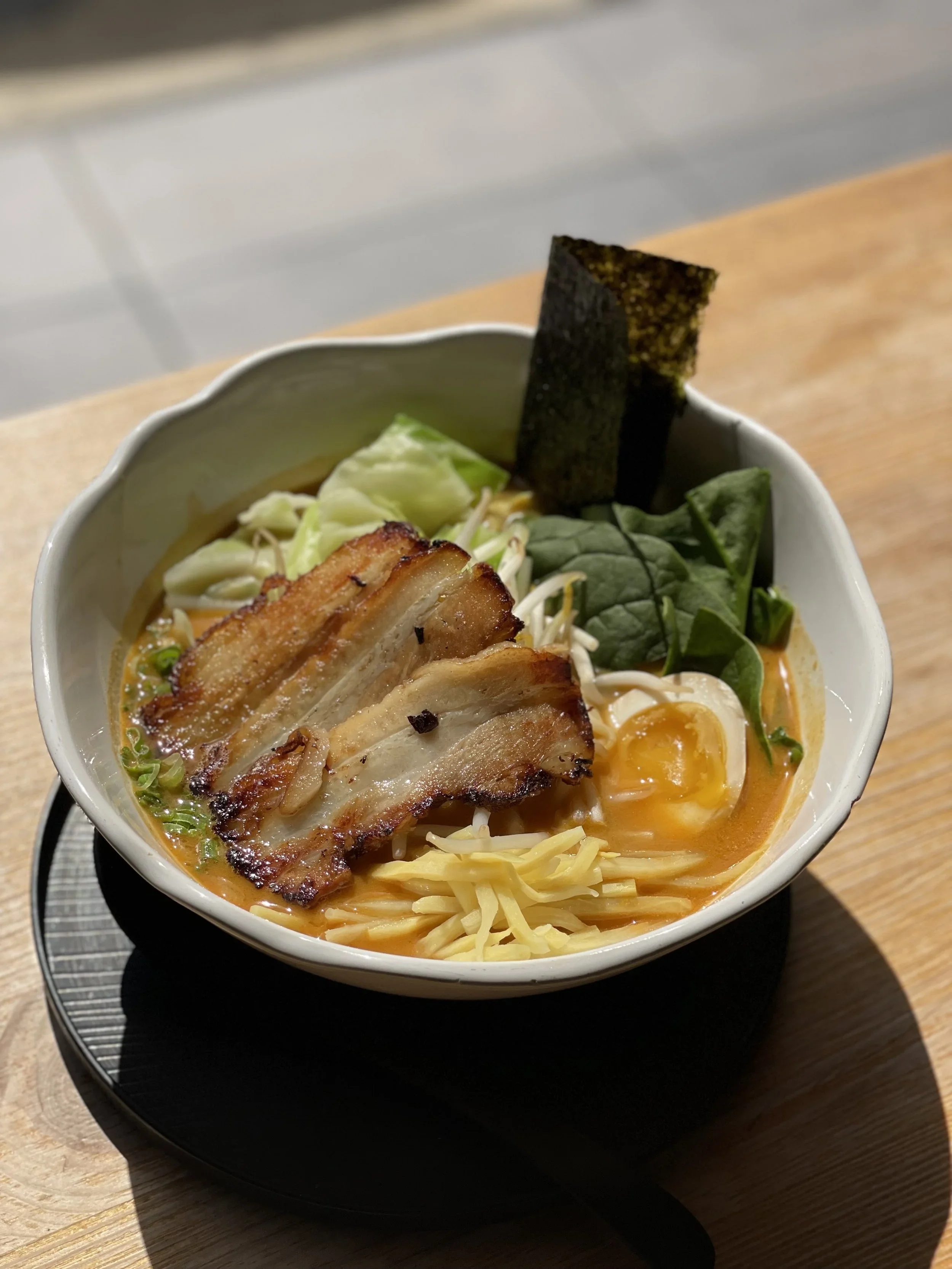 A bowl of ramen noodles with crispy pork belly, soft-boiled egg, seaweed, spinach, bean sprouts, and shredded cheese on top, served on a black tray on a wooden table.