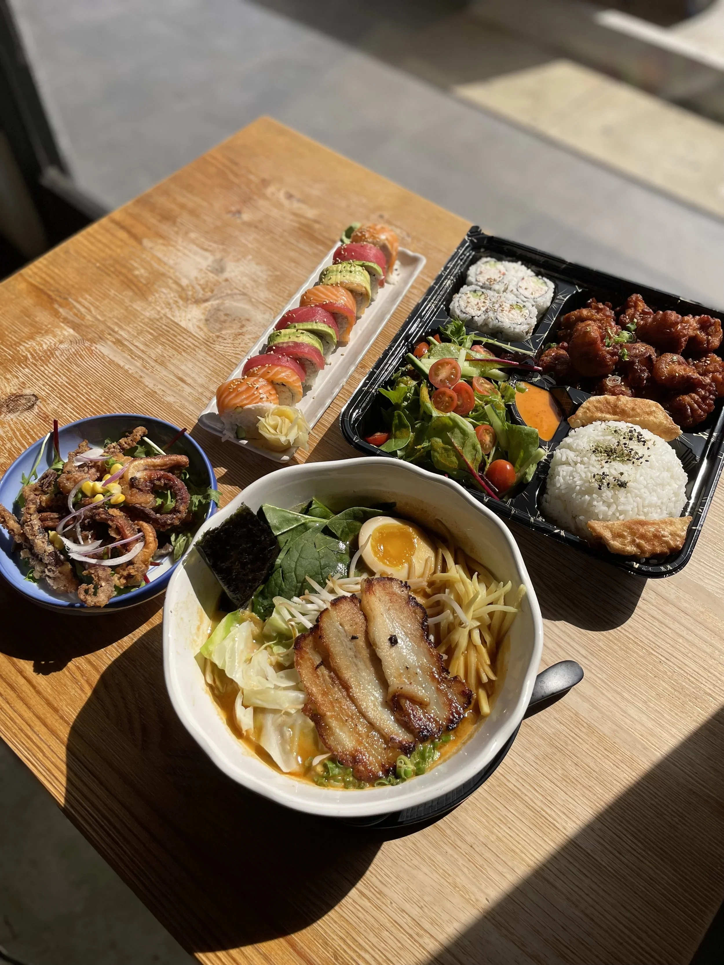 A variety of Asian dishes on a wooden table, including sushi rolls, a salad with cherry tomatoes, fried chicken, a rice bowl with bacon and greens, and an octopus dish.